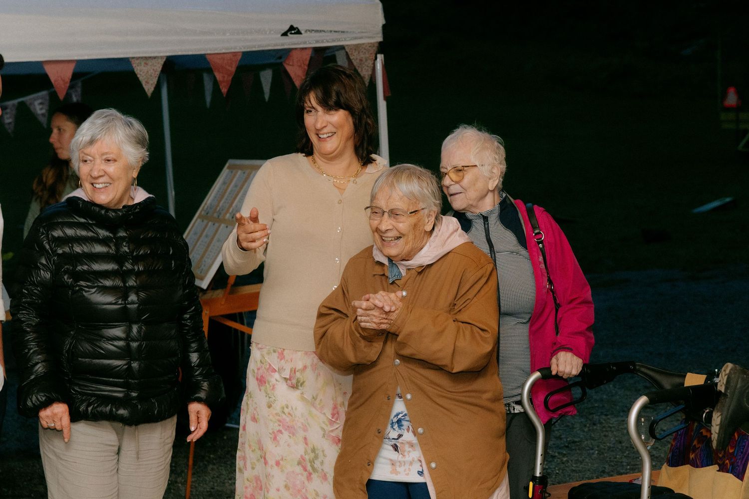 Group of elderly women smiling and clapping outdoors at a community event with bunting decorations.