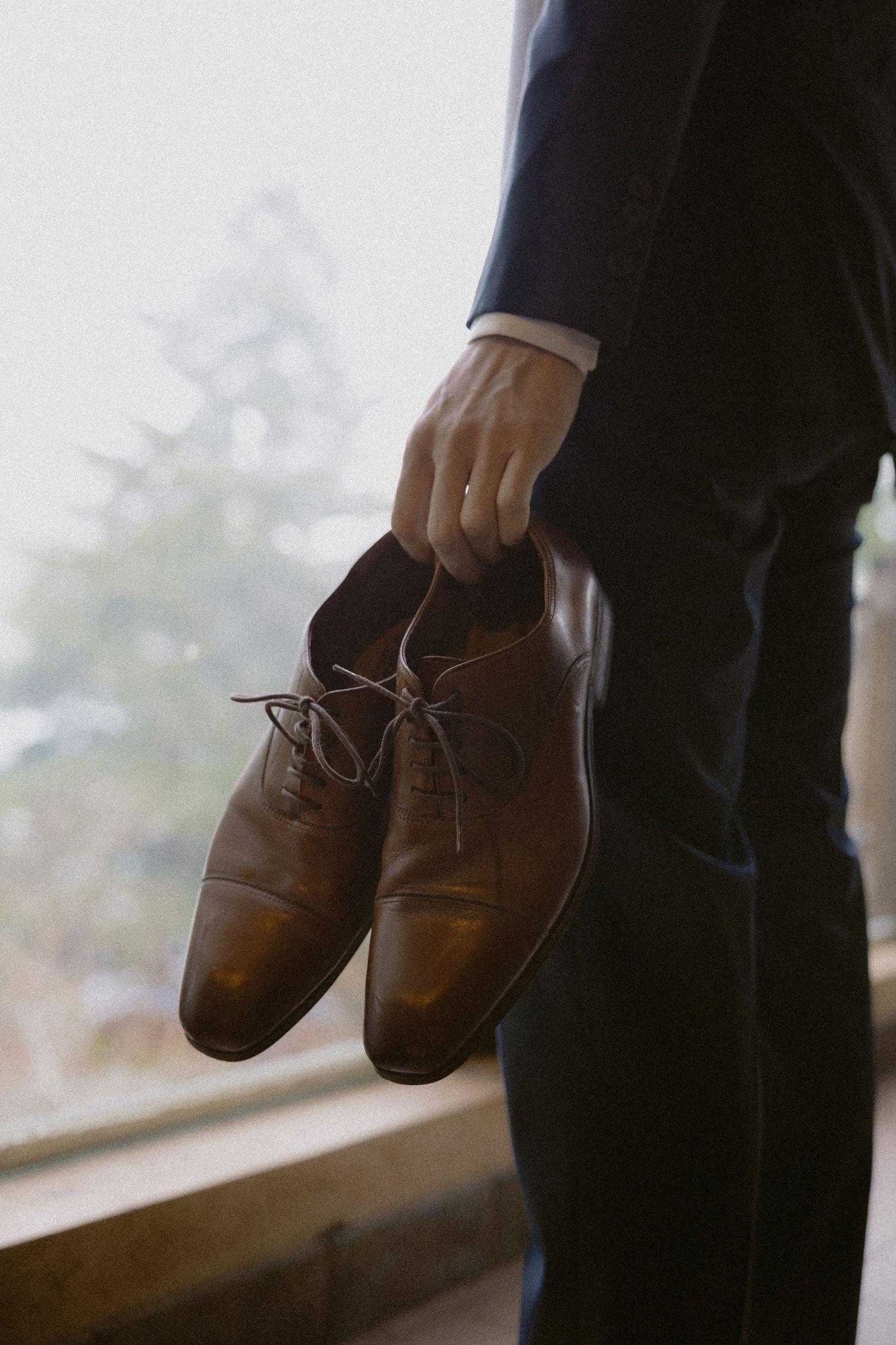 Brown leather dress shoes being held up against a window with natural lighting.
