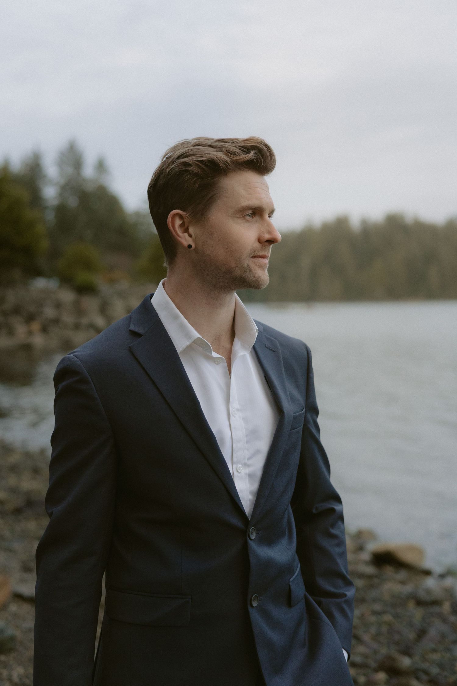A person in a navy suit and white shirt gazes thoughtfully across a serene lake landscape.