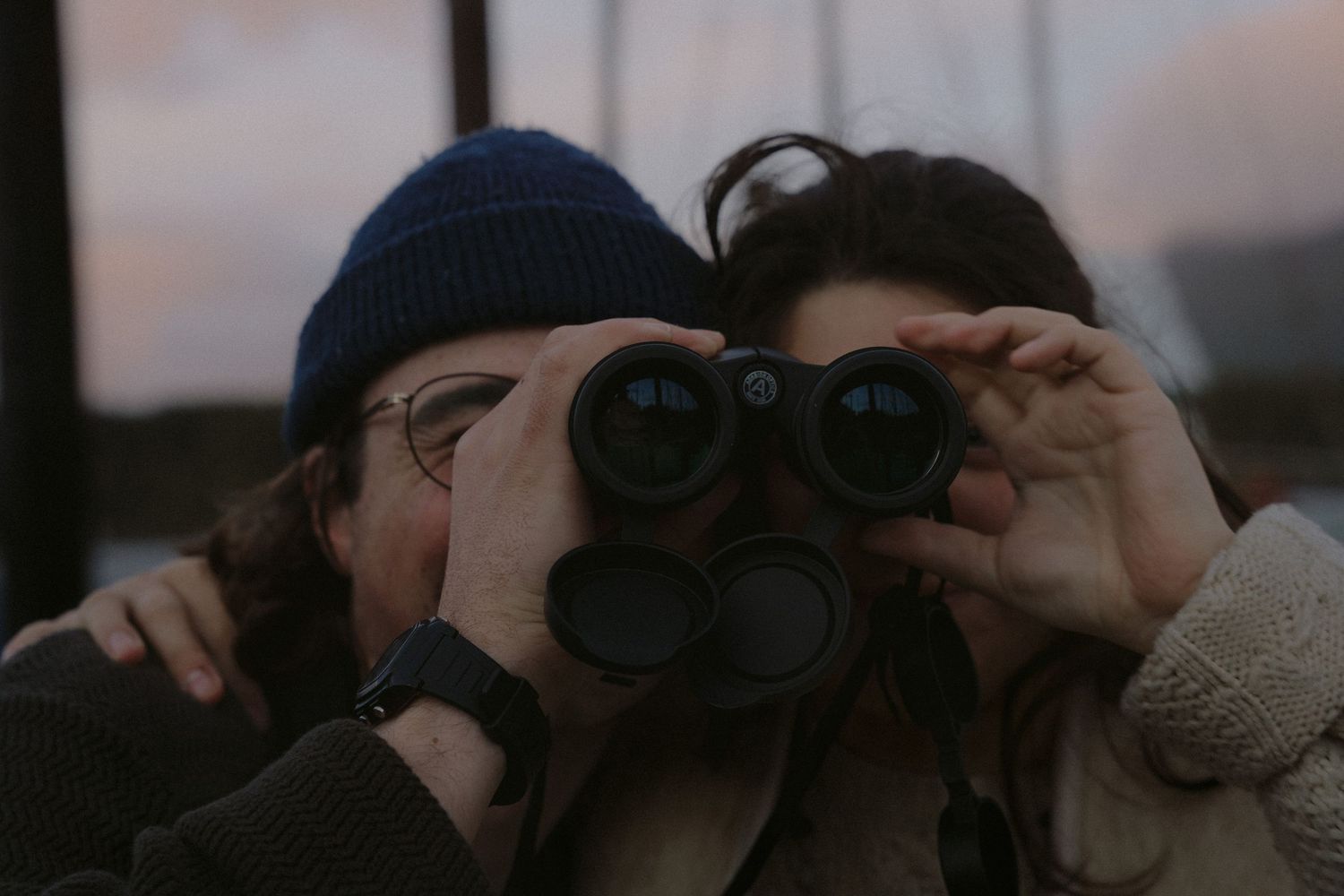 Person using binoculars to observe from waterfront at sunset.