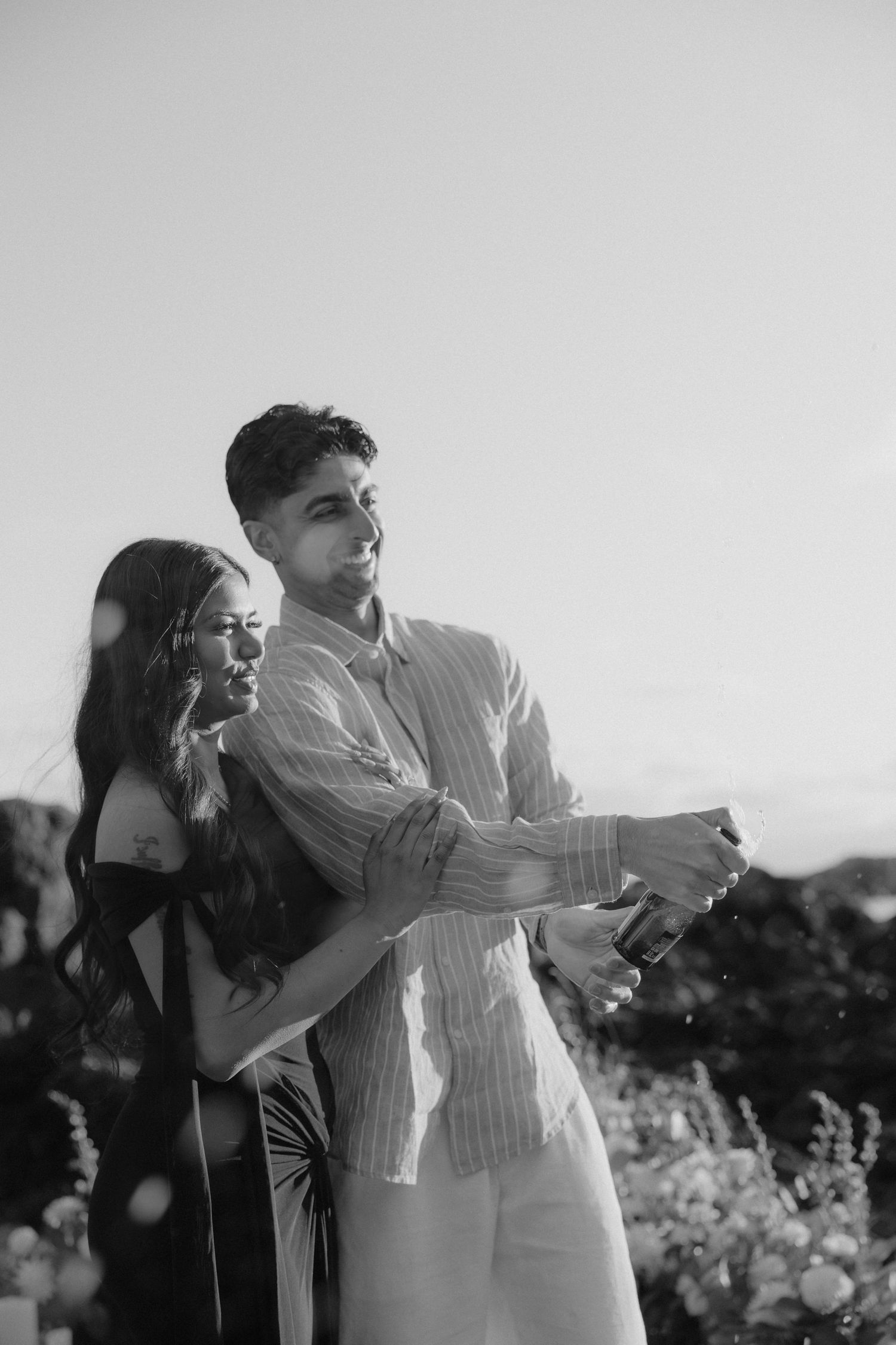 Black and white photo of couple popping champagne bottle outdoors at sunset.