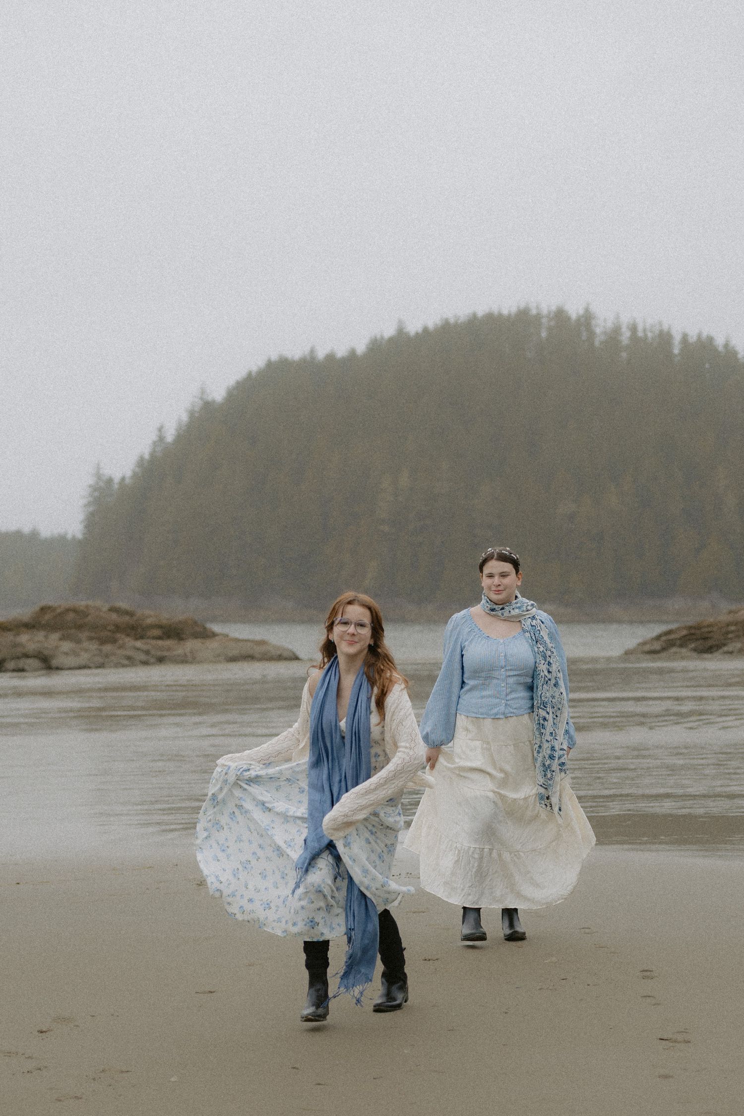 Two people in denim jackets and wedding attire walking along a misty beach with evergreen-covered islands.