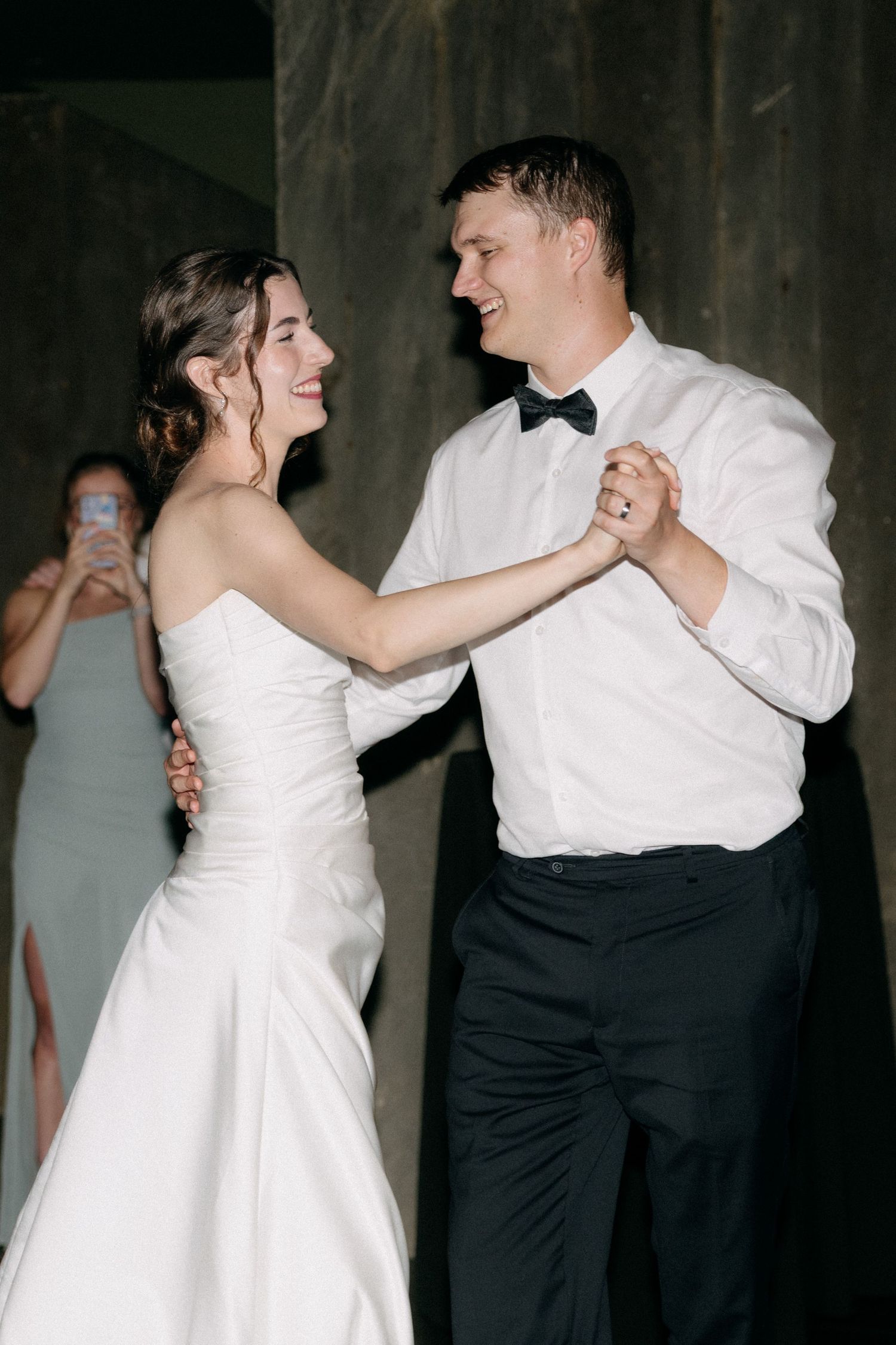 Bride and groom share their first dance, smiling joyfully, as guests capture the moment in the background.