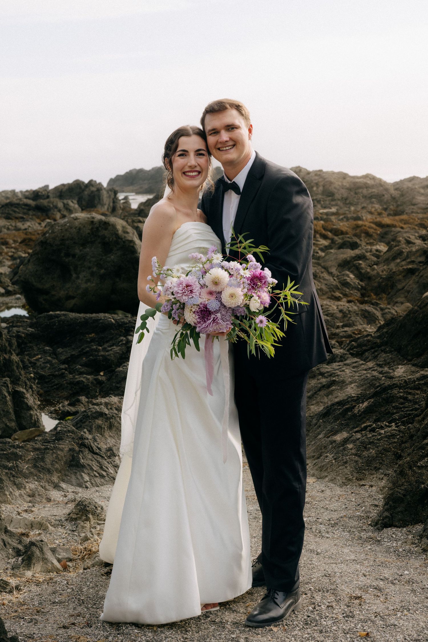 Bride and groom pose together on rocky coastal shore, she in white gown holding purple floral bouquet, he in black tuxedo.