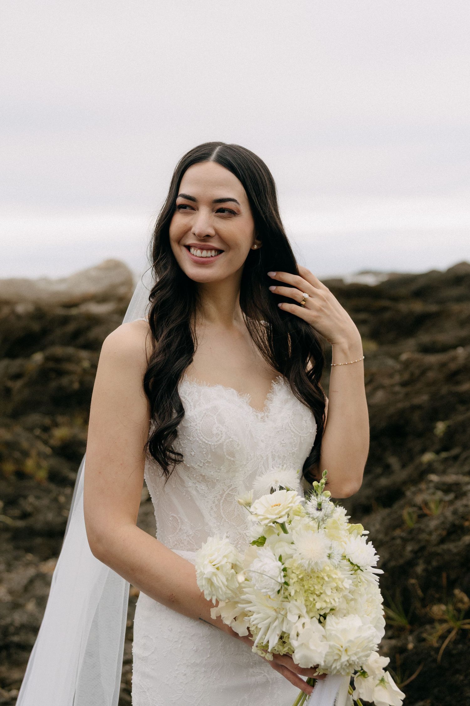 Smiling bride in white lace gown holds white floral bouquet outdoors near rocky coast.