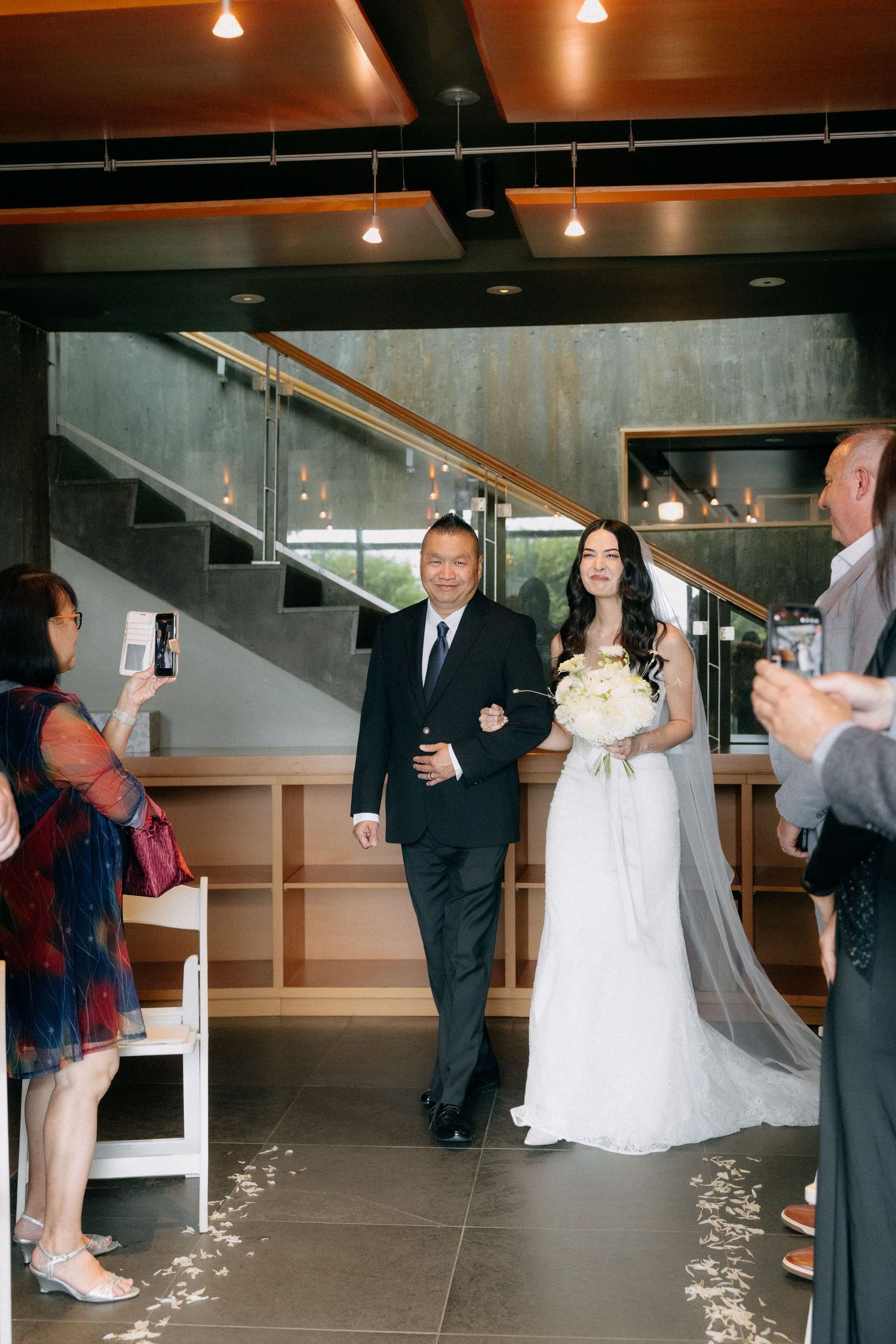 Bride in white gown walks down the aisle with her father during an indoor wedding ceremony.