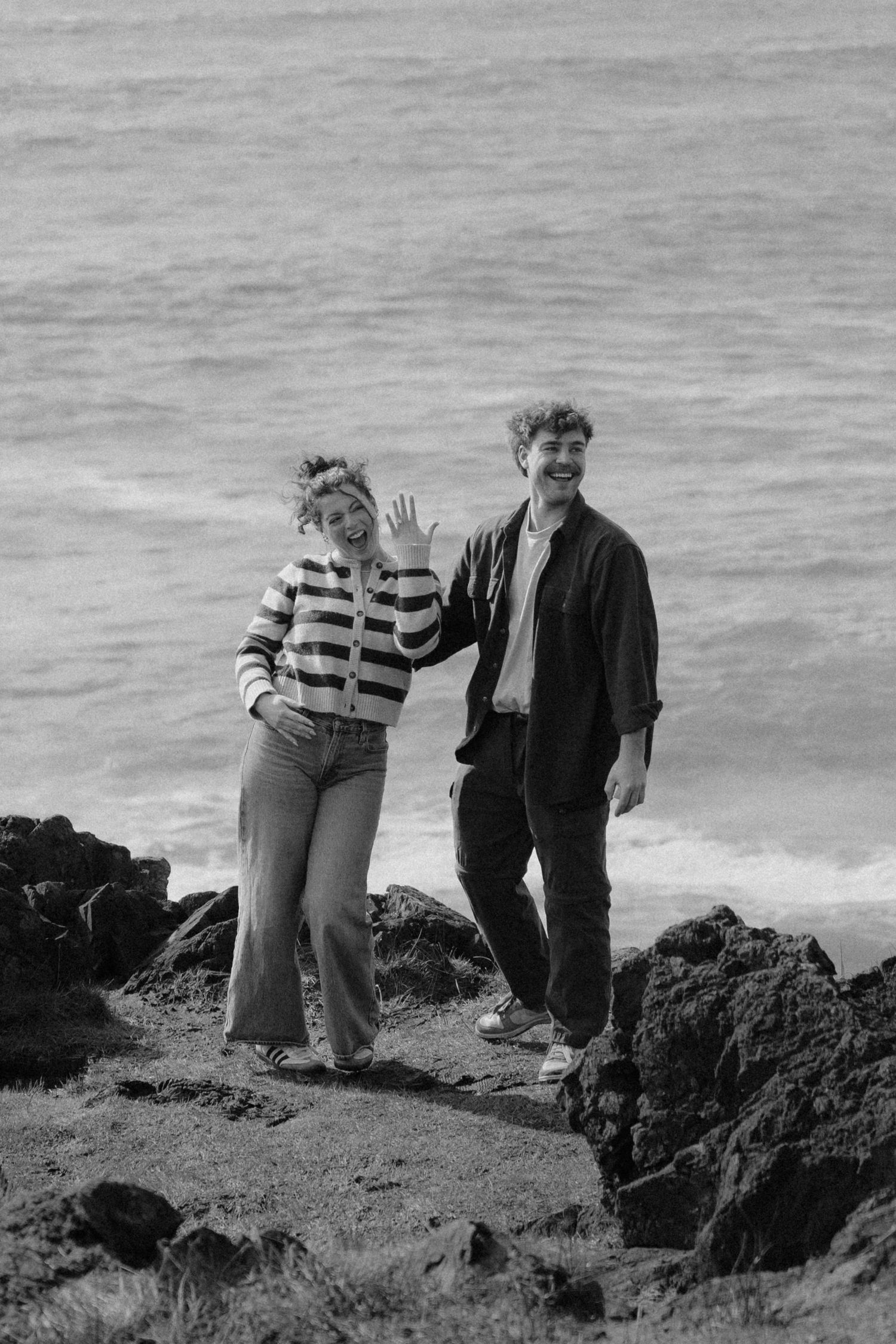 Happy couple waving and smiling together on rocky coastal cliffs in black and white photo.