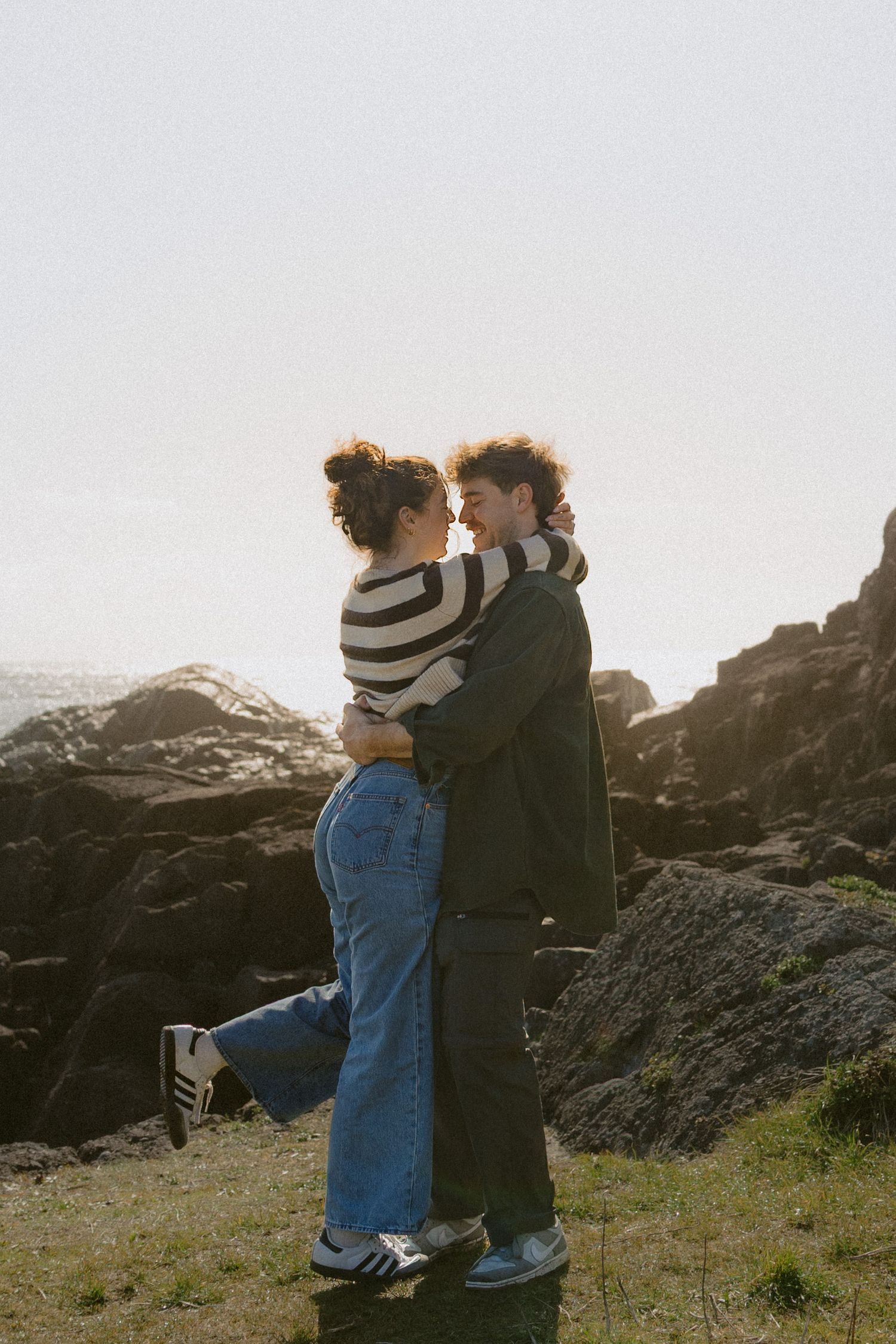 Romantic couple embracing and smiling face to face on coastal cliffs during golden hour.