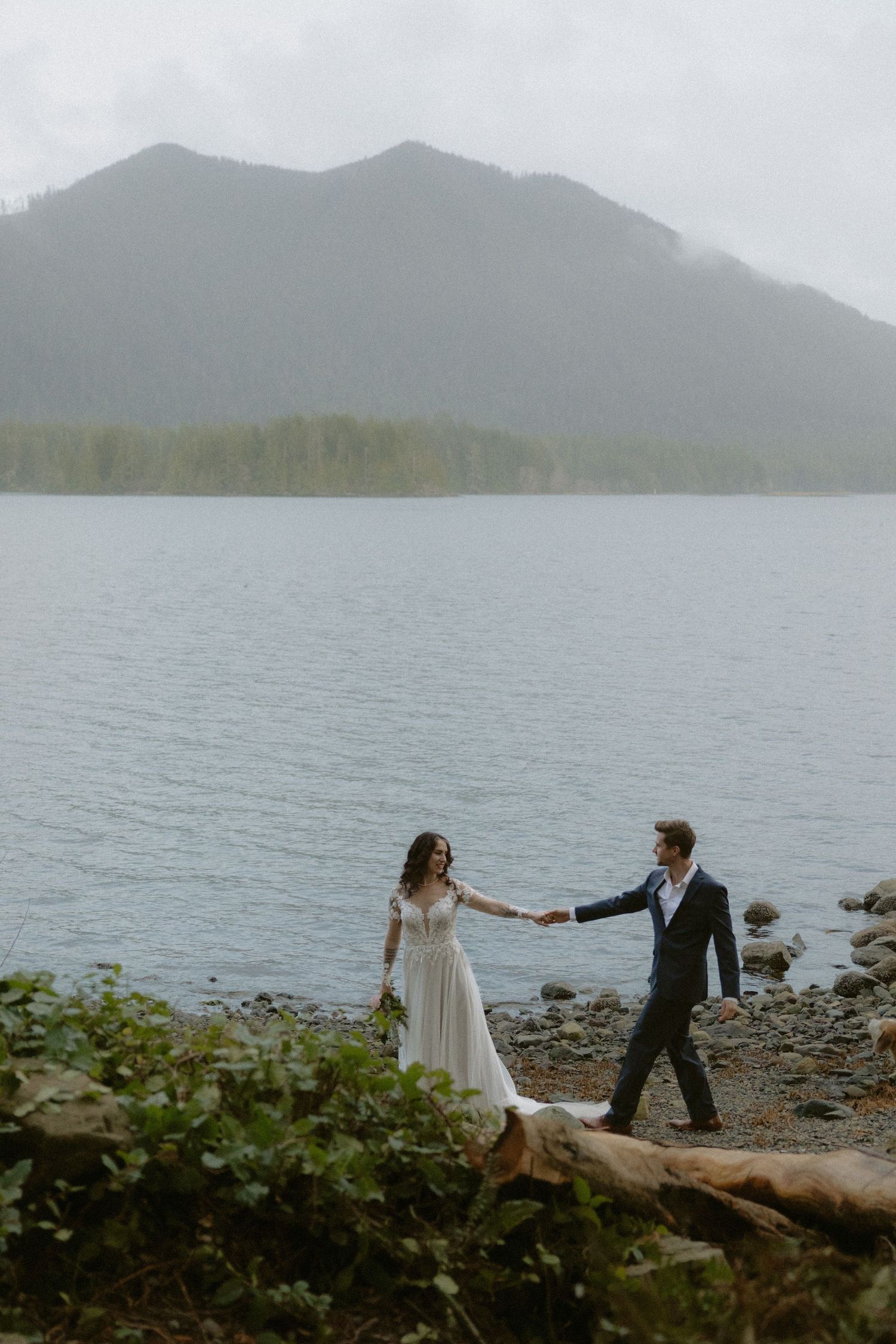 A couple dances together on a rocky beach with mountains and evergreen trees creating a dramatic backdrop.