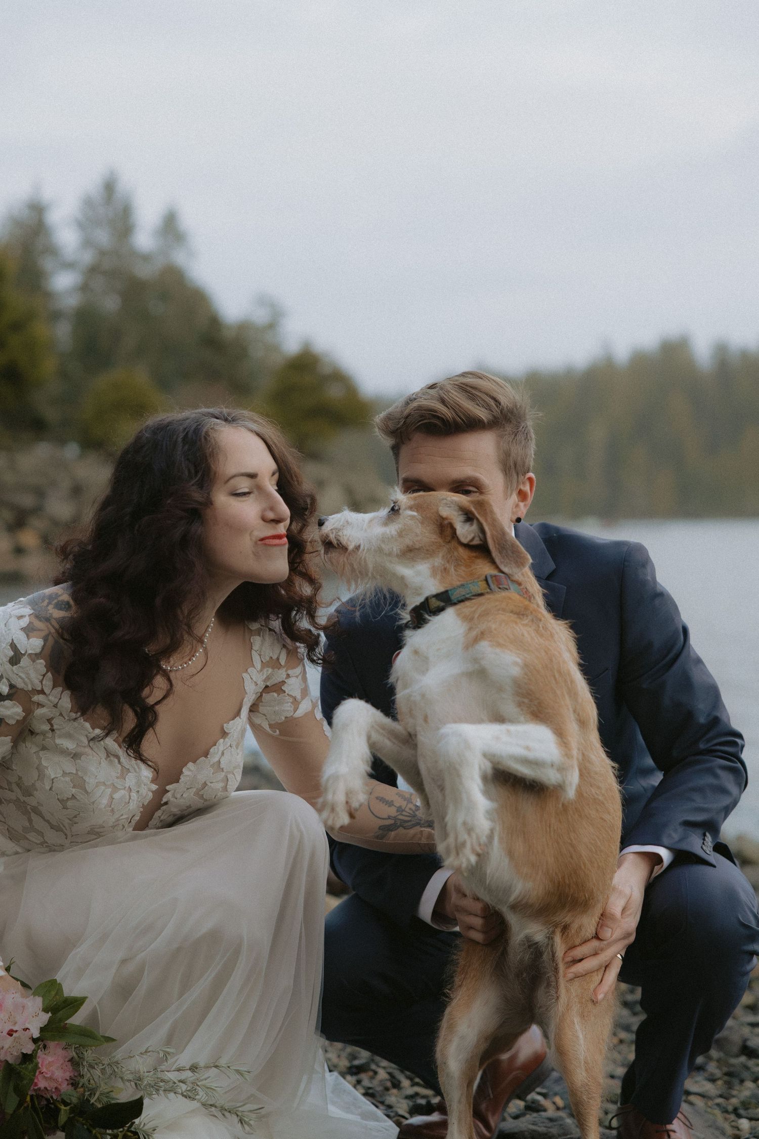 A couple plays with their dog during a wedding photoshoot on a rocky beach.