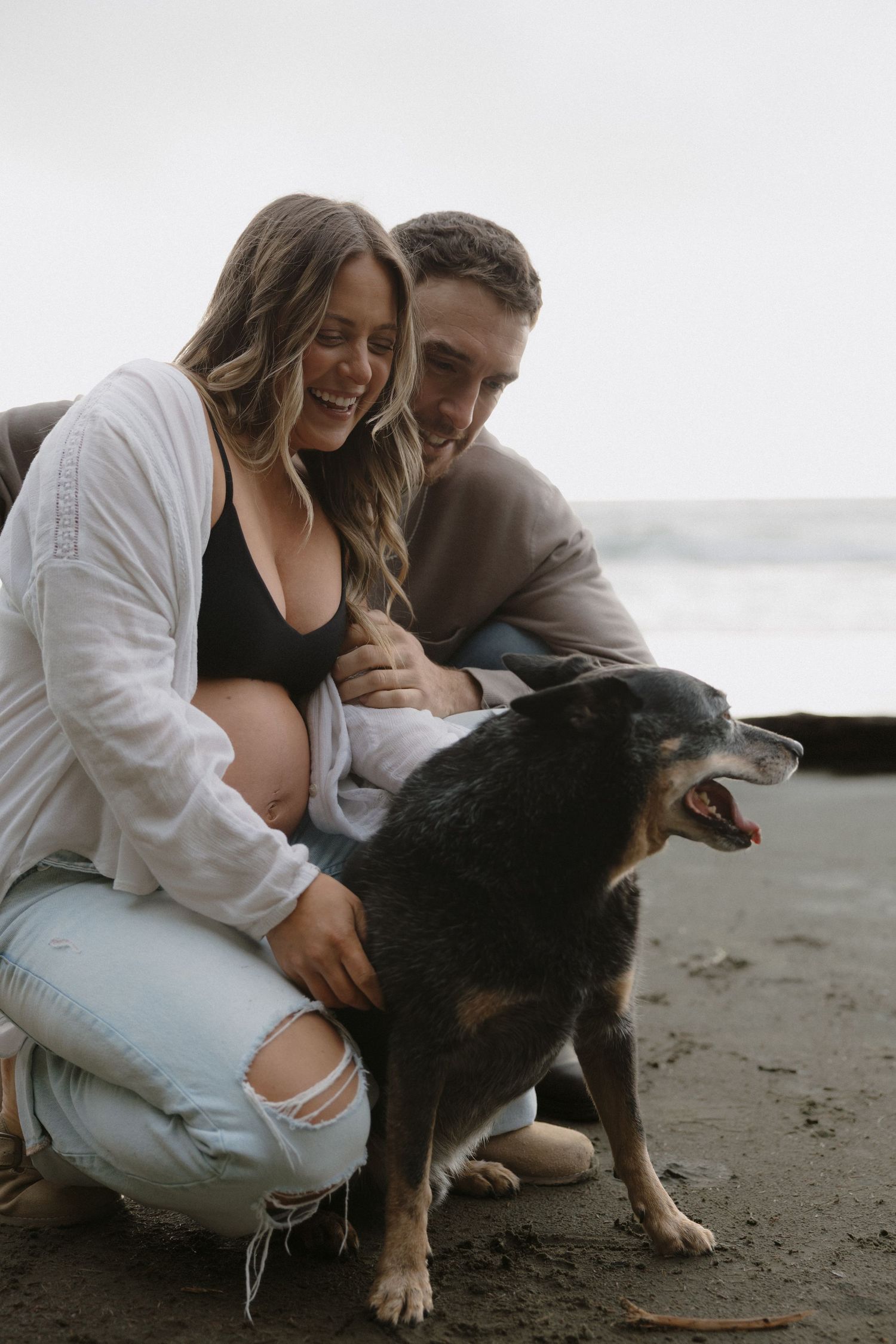 A couple shares tender moments with their black dog on a cloudy beach.