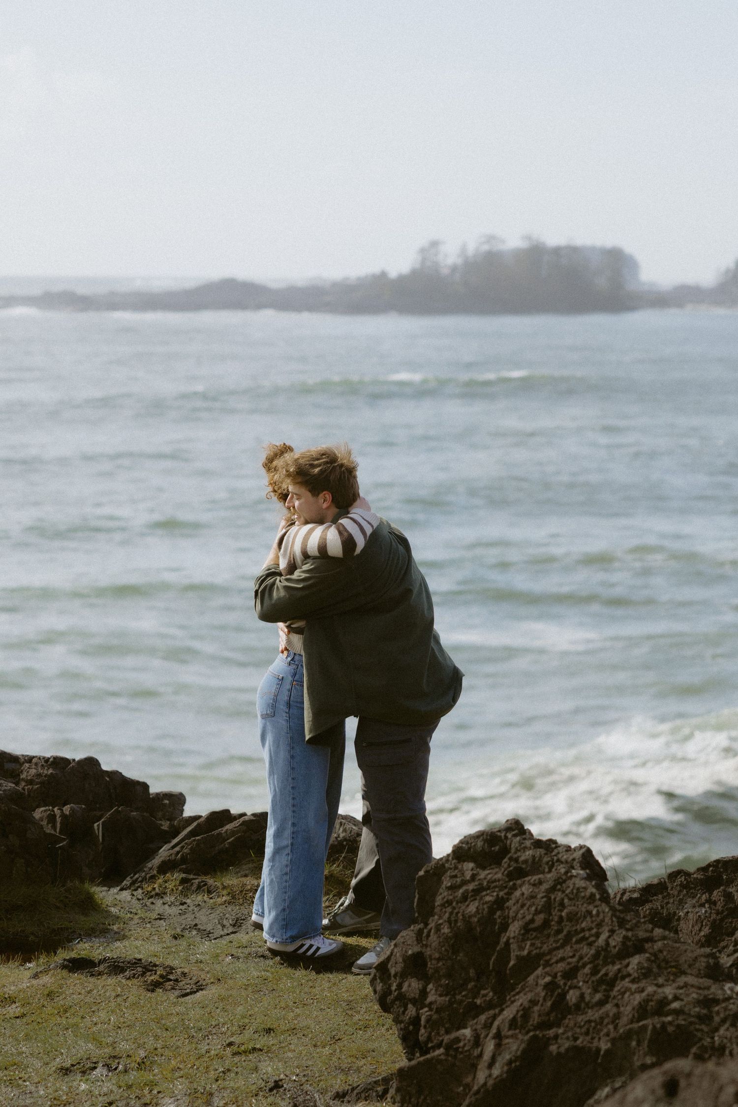 Newly engaged couple embracing tenderly on rocky beach with calm ocean and island in background.