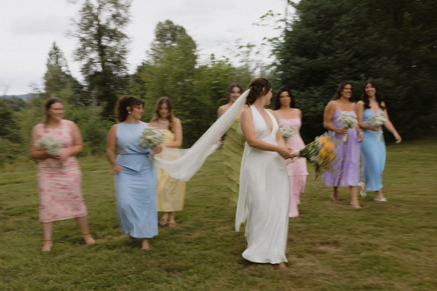 Bride in white gown kicks leg playfully with bridesmaids in colorful pastel dresses on green lawn.