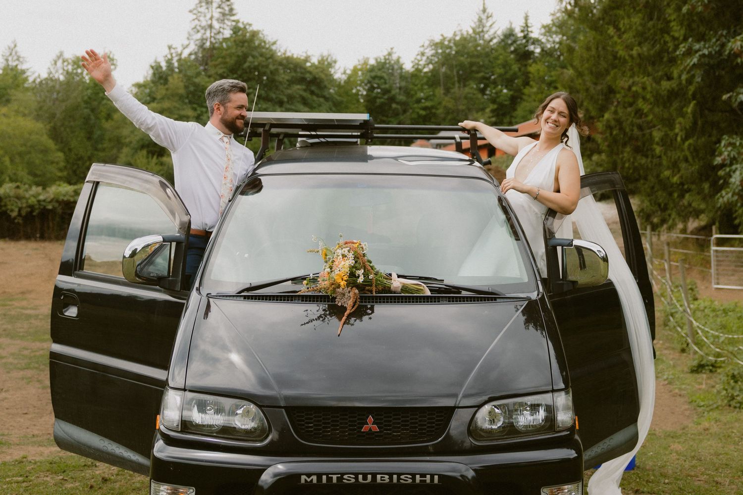 Bride and groom celebrating beside a decorated black Mitsubishi SUV with floral bouquet at outdoor wedding.