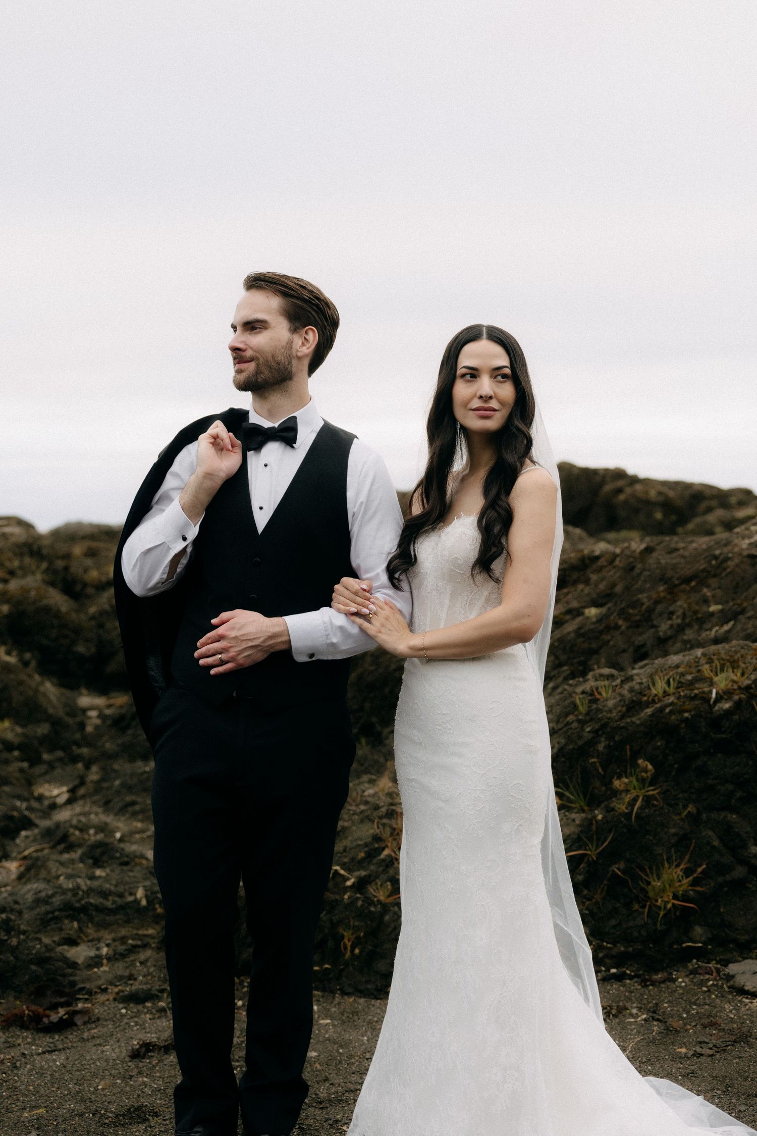 Bride in white gown and groom in black tuxedo pose together on rocky coastal landscape under overcast sky.