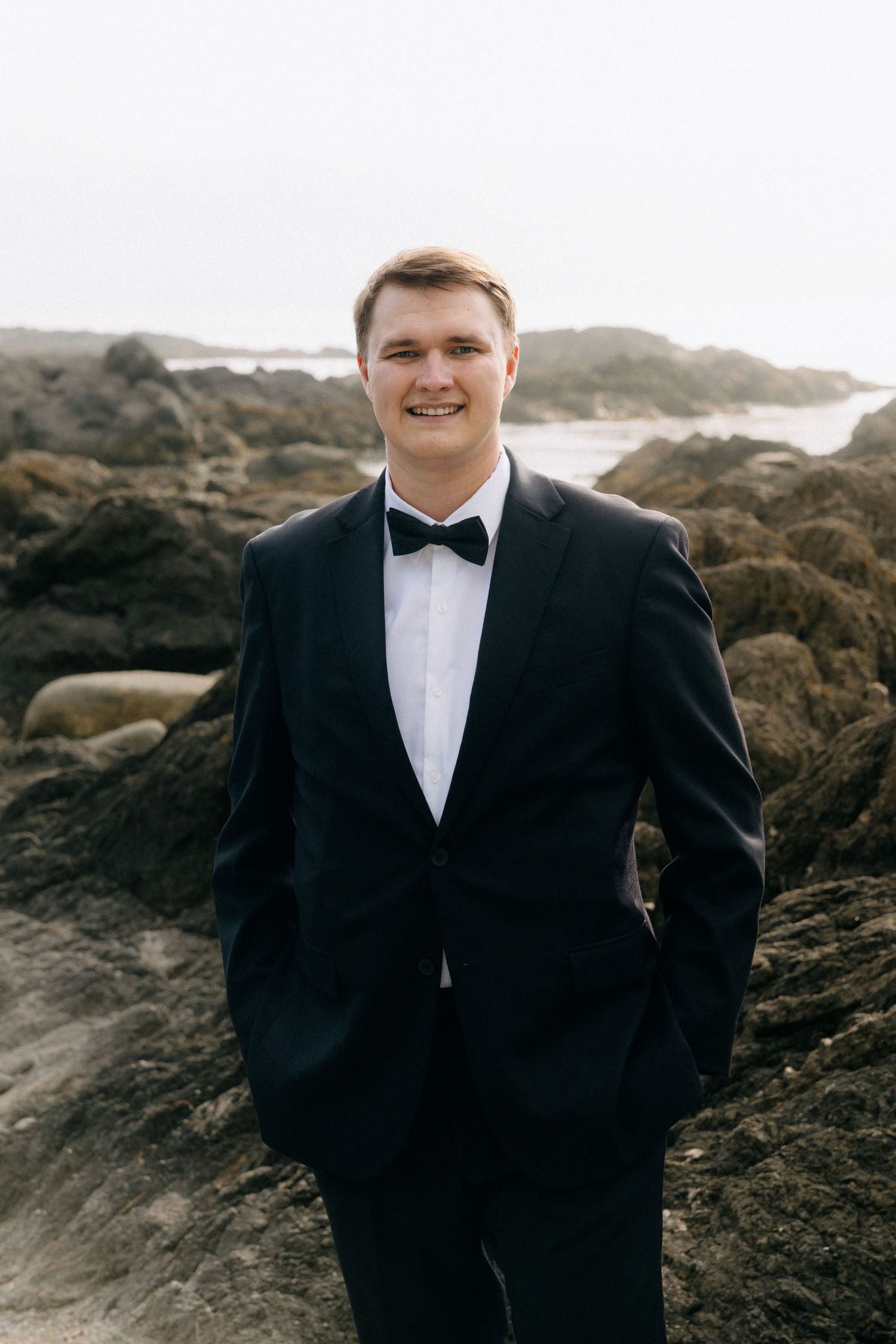 Smiling groom in black tuxedo with bow tie standing on rocky coastal shoreline.