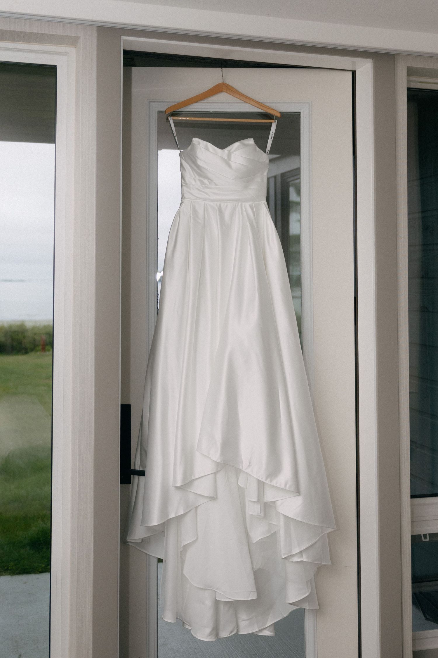 White strapless ball gown wedding dress with layered skirt hanging on wooden hanger in front of glass door.