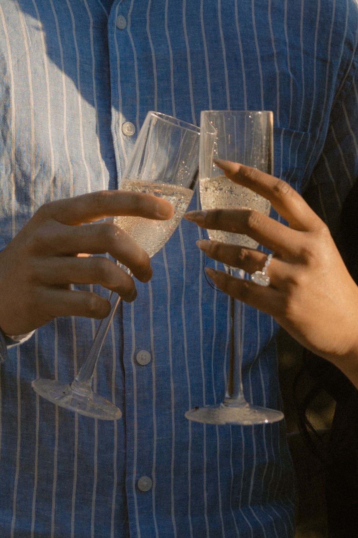Two hands clinking champagne flutes in a celebratory toast, held by a person in a blue striped shirt.