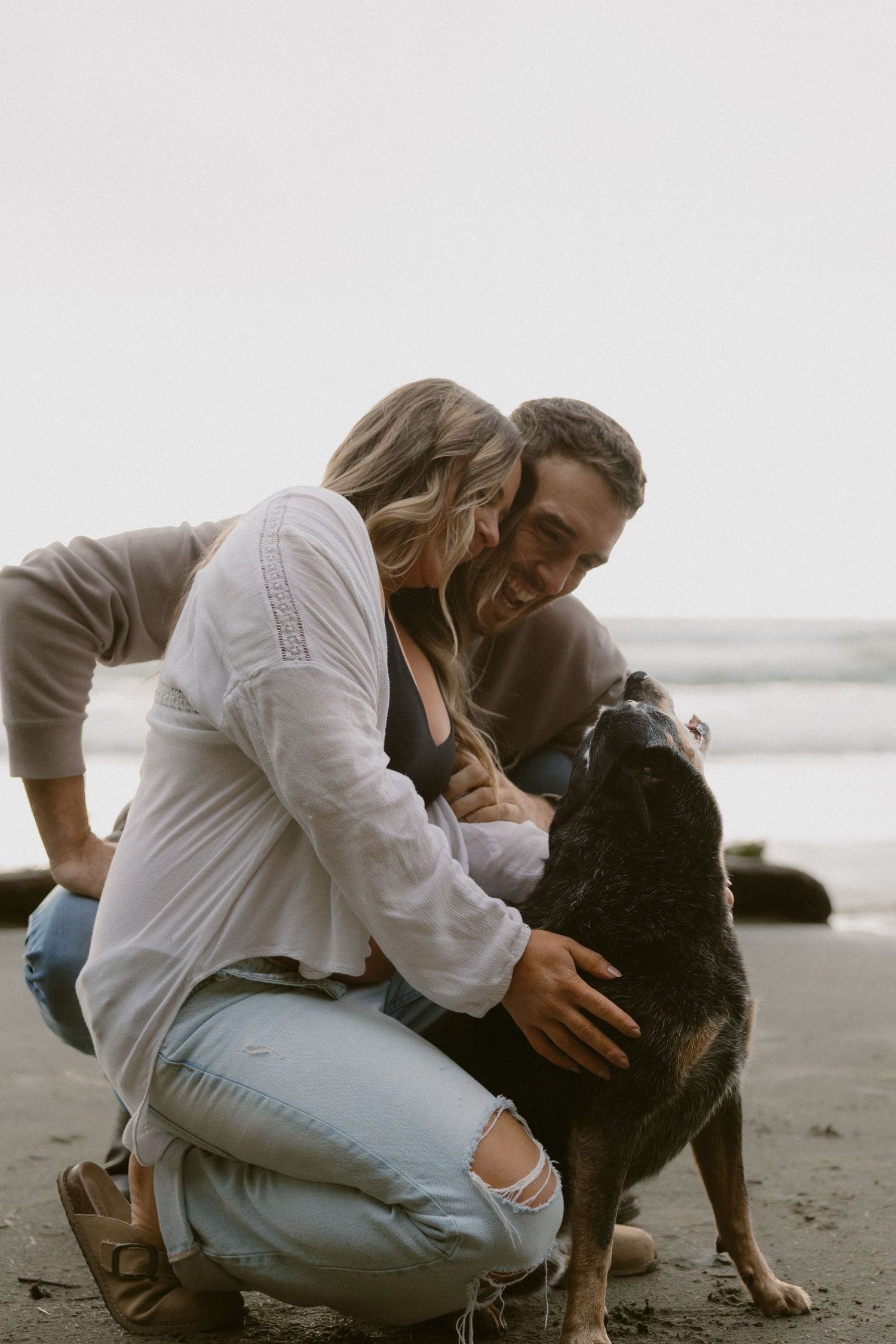 A couple sits on a beach with their black dog during an intimate photoshoot.