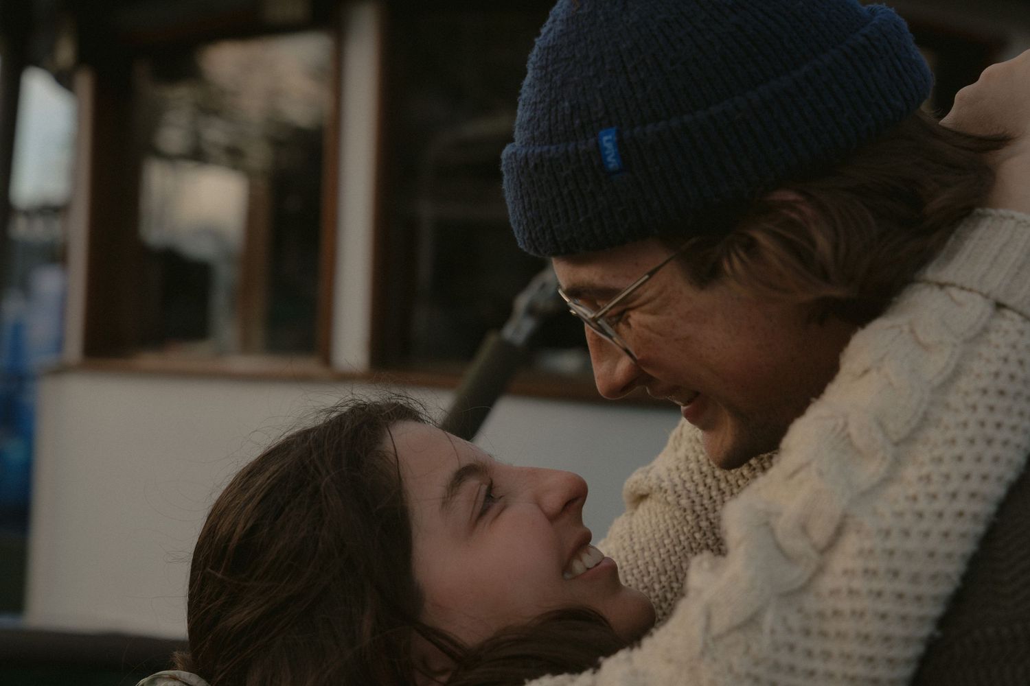 A close embrace between two people with boats and marina structures visible in the background.