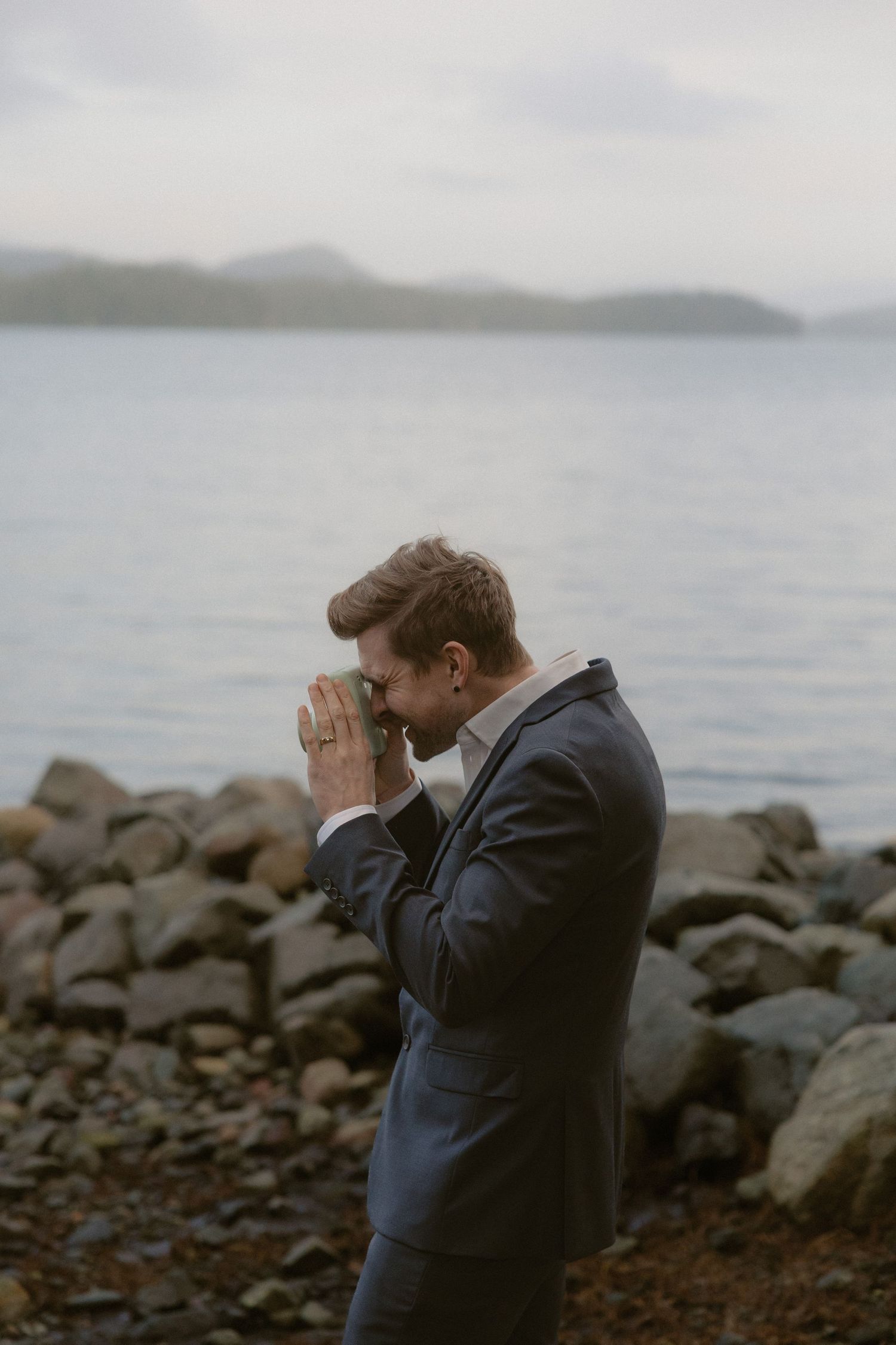 A person in a navy suit stands by the rocky shoreline looking out over the misty water.