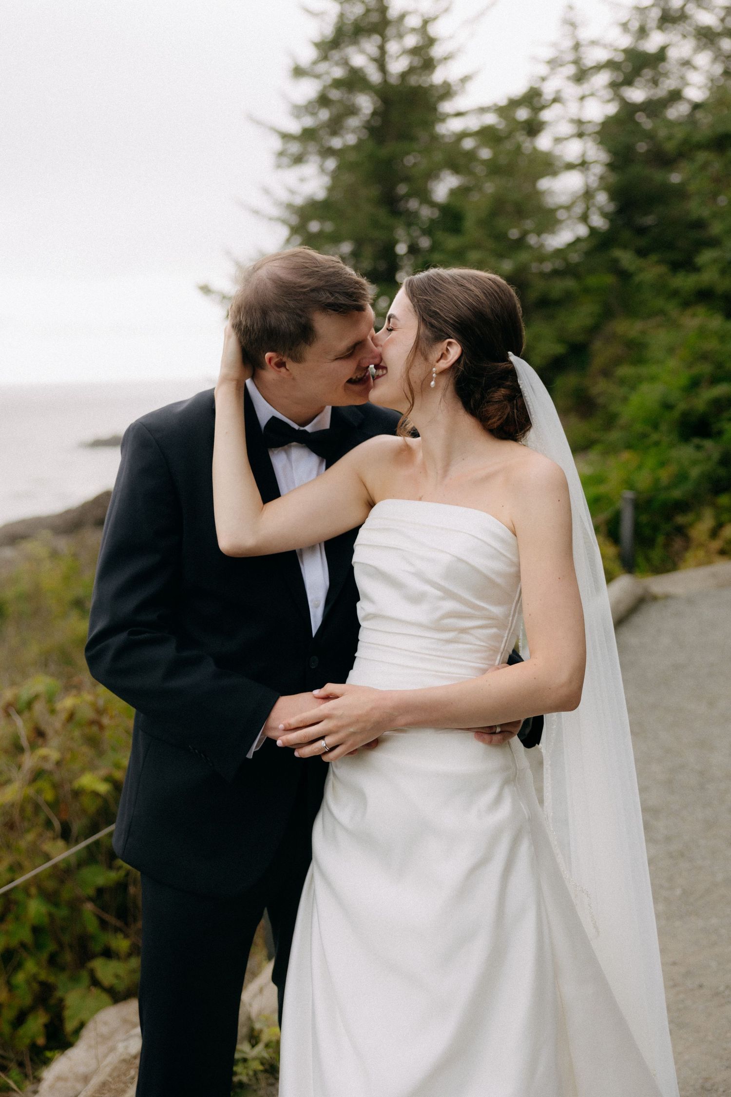 Bride in strapless white gown with veil and groom in black tuxedo sharing a tender kiss outdoors.