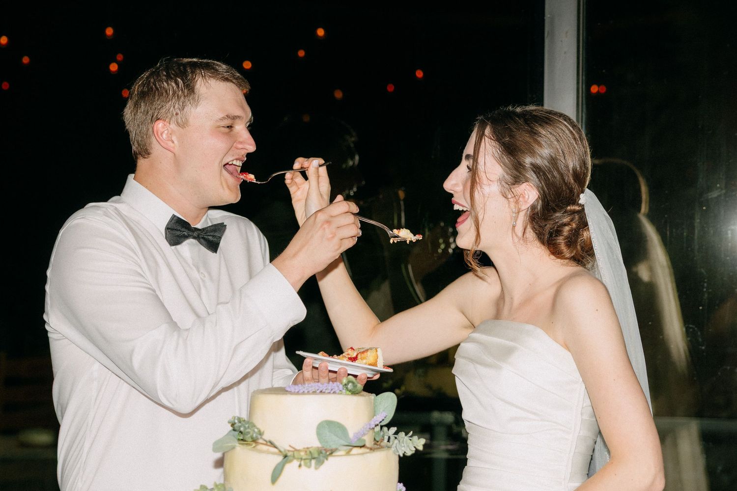 Bride and groom laughing while feeding each other wedding cake at their reception.