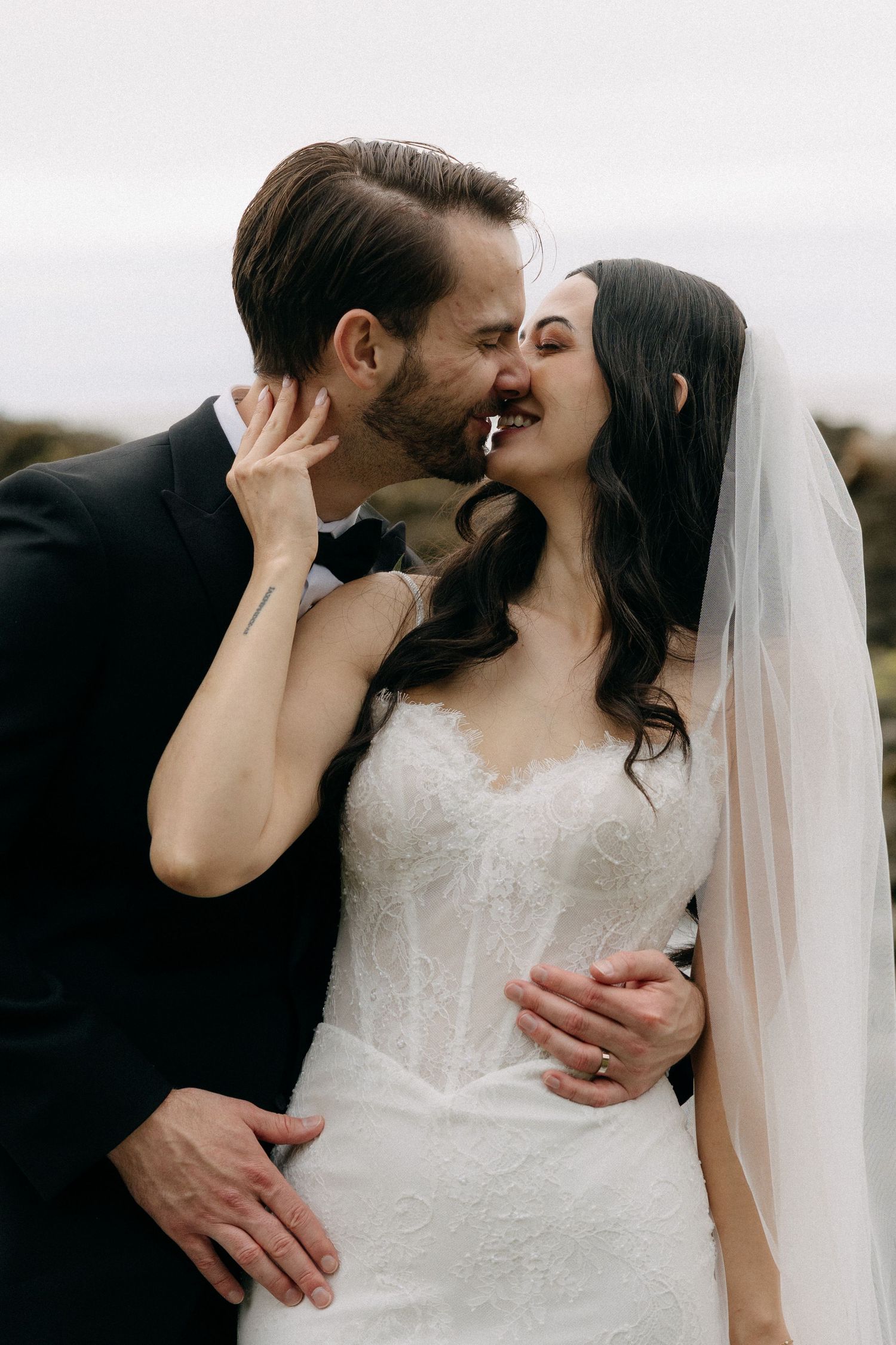 A groom in a black suit tenderly kisses a smiling bride in a white lace gown and veil outdoors.