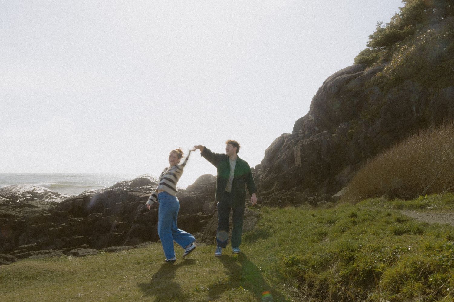Joyful couple dancing and spinning hands on green coastal hillside with rocky cliffs behind.