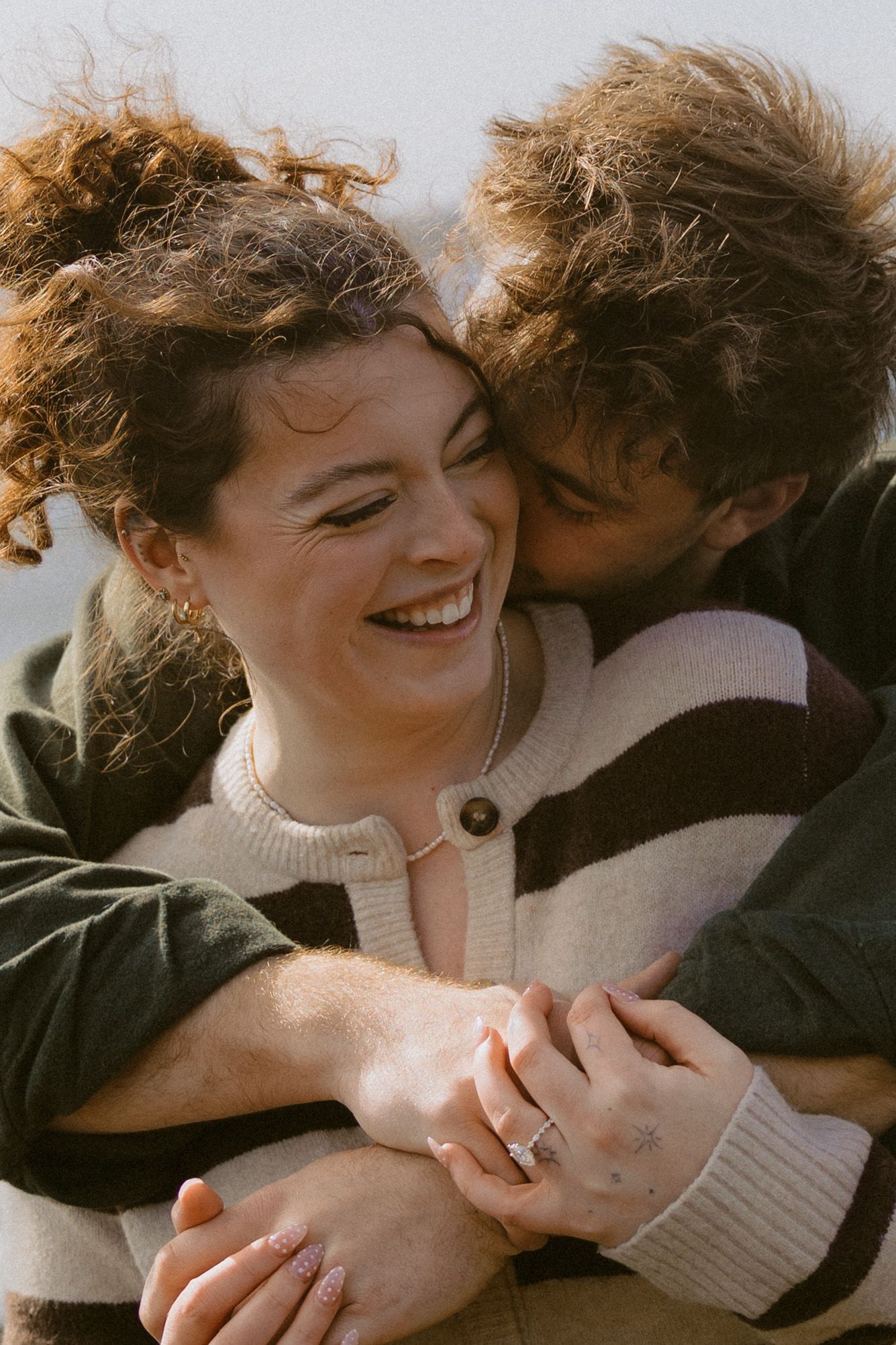 Happy couple embracing on beach, woman laughing as man kisses her cheek, showing engagement ring.