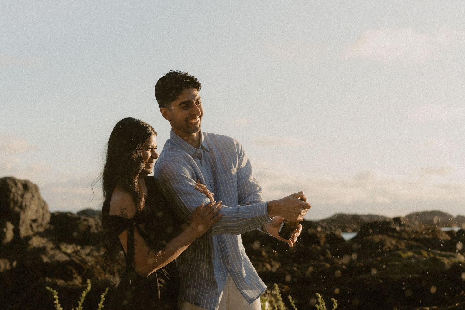 Couple dancing and laughing together on rocky coastal landscape during golden hour sunset.