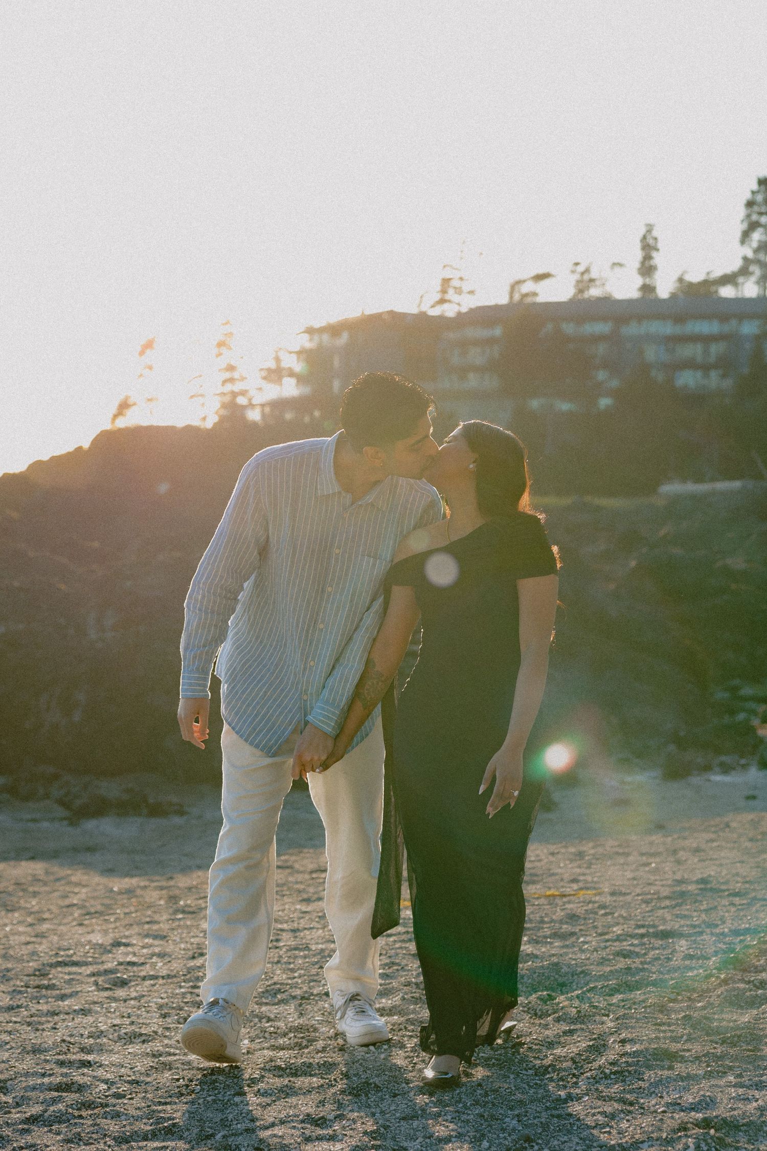 Couple sharing a kiss while walking on rocky terrain during a golden sunset outdoors.