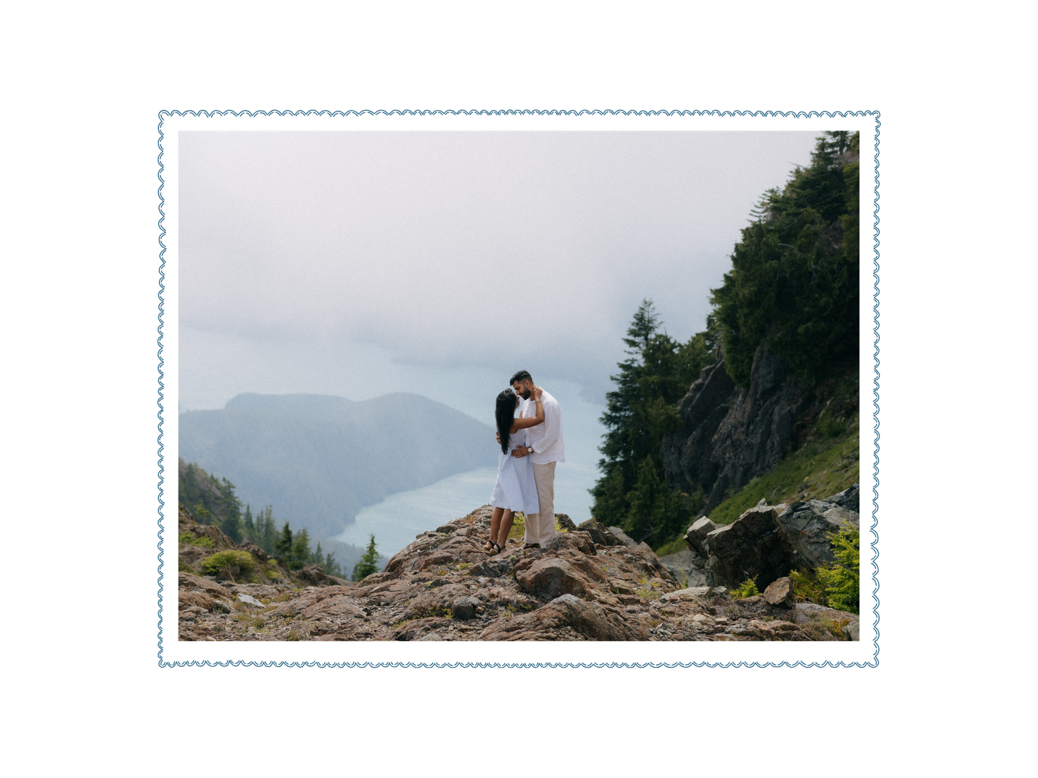 Couple embracing on rocky mountain summit with misty fjord and evergreen trees in background.