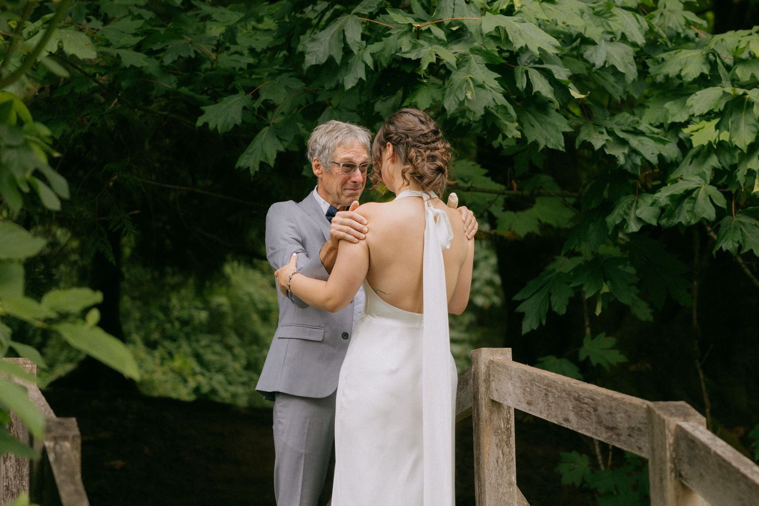 Bride in backless wedding gown shares an emotional first look moment with an older gentleman on a wooden bridge.
