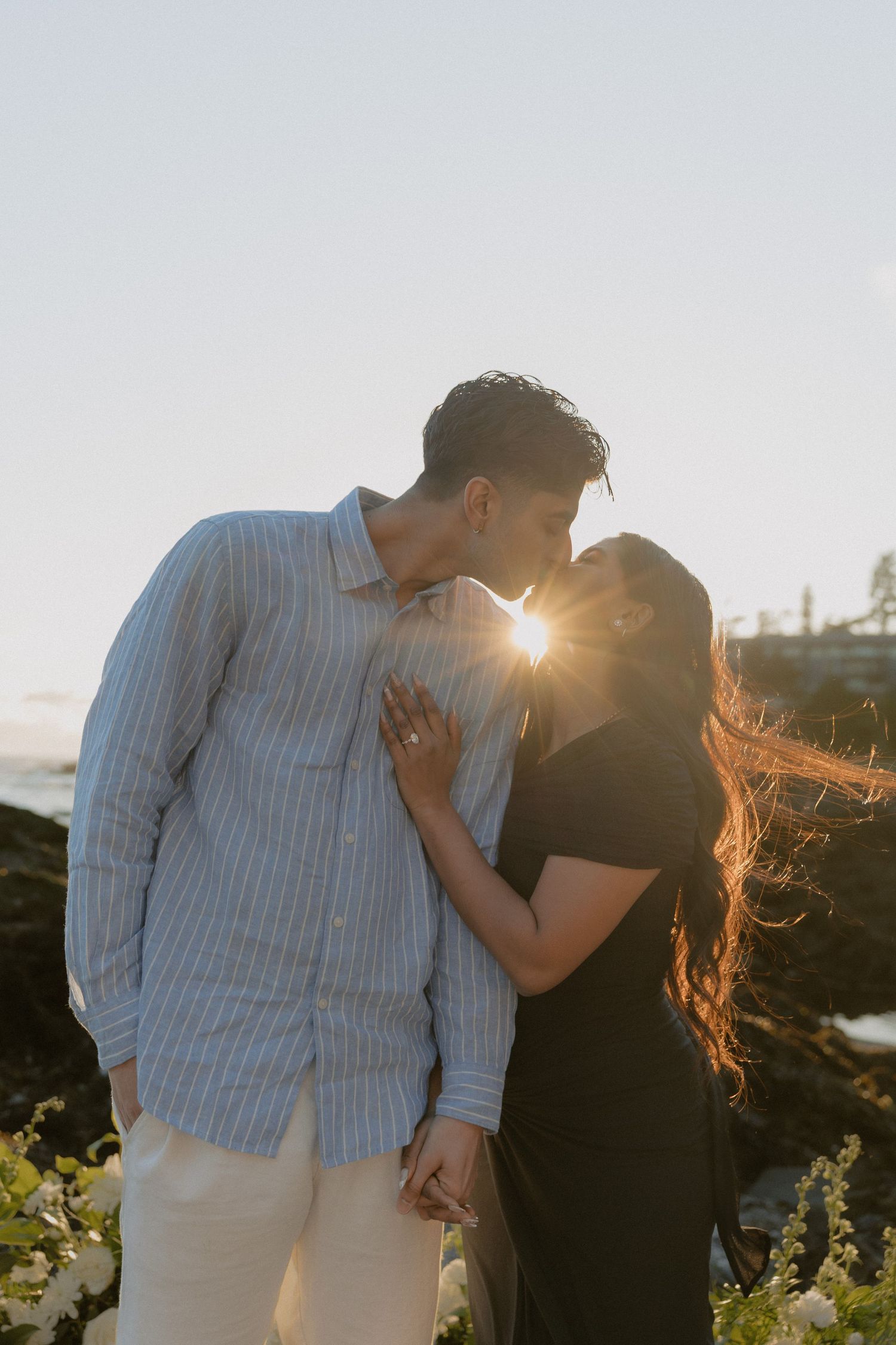 Couple kissing at golden hour on rocky shoreline, sun flare between them, white flowers in foreground.