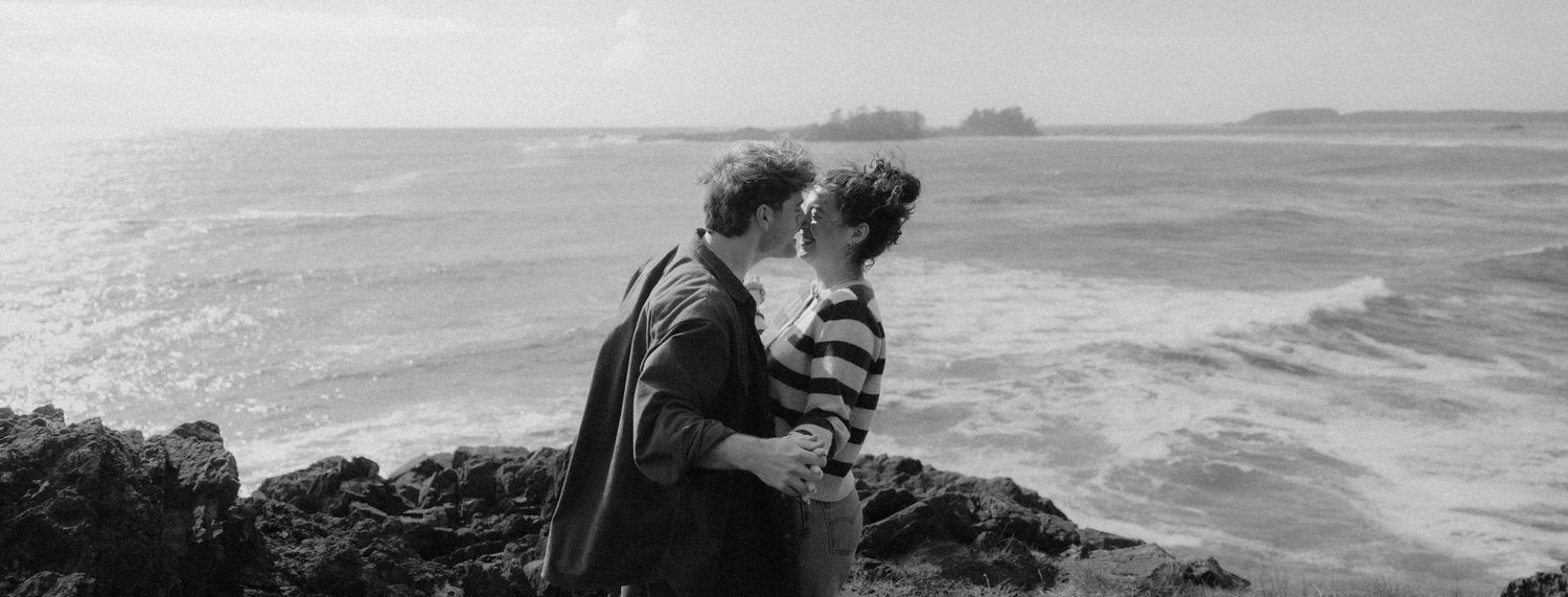 Black and white photo of couple embracing and kissing on rocky ocean cliffs with waves behind them.