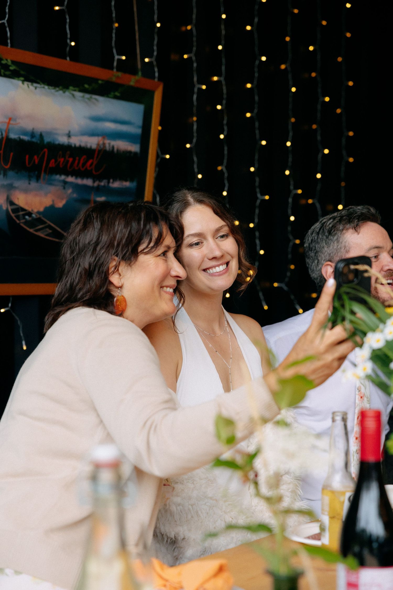 Bride in white dress smiling at phone with guests at wedding reception table, fairy lights and married sign in background.