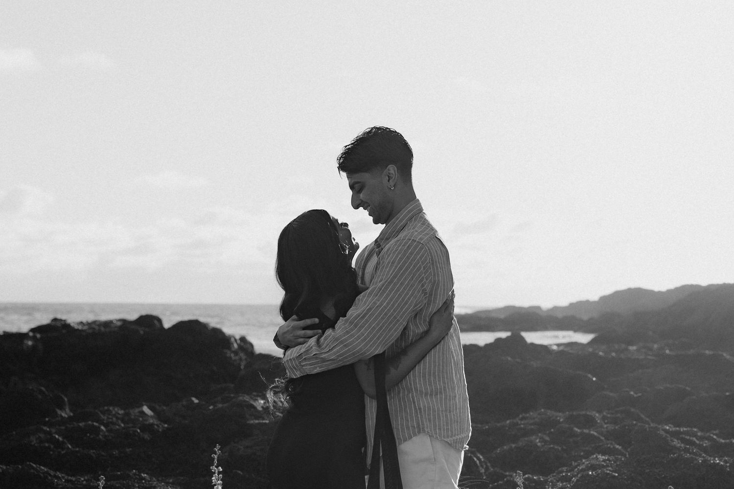 Couple embracing romantically on rocky ocean coastline in black and white photo.