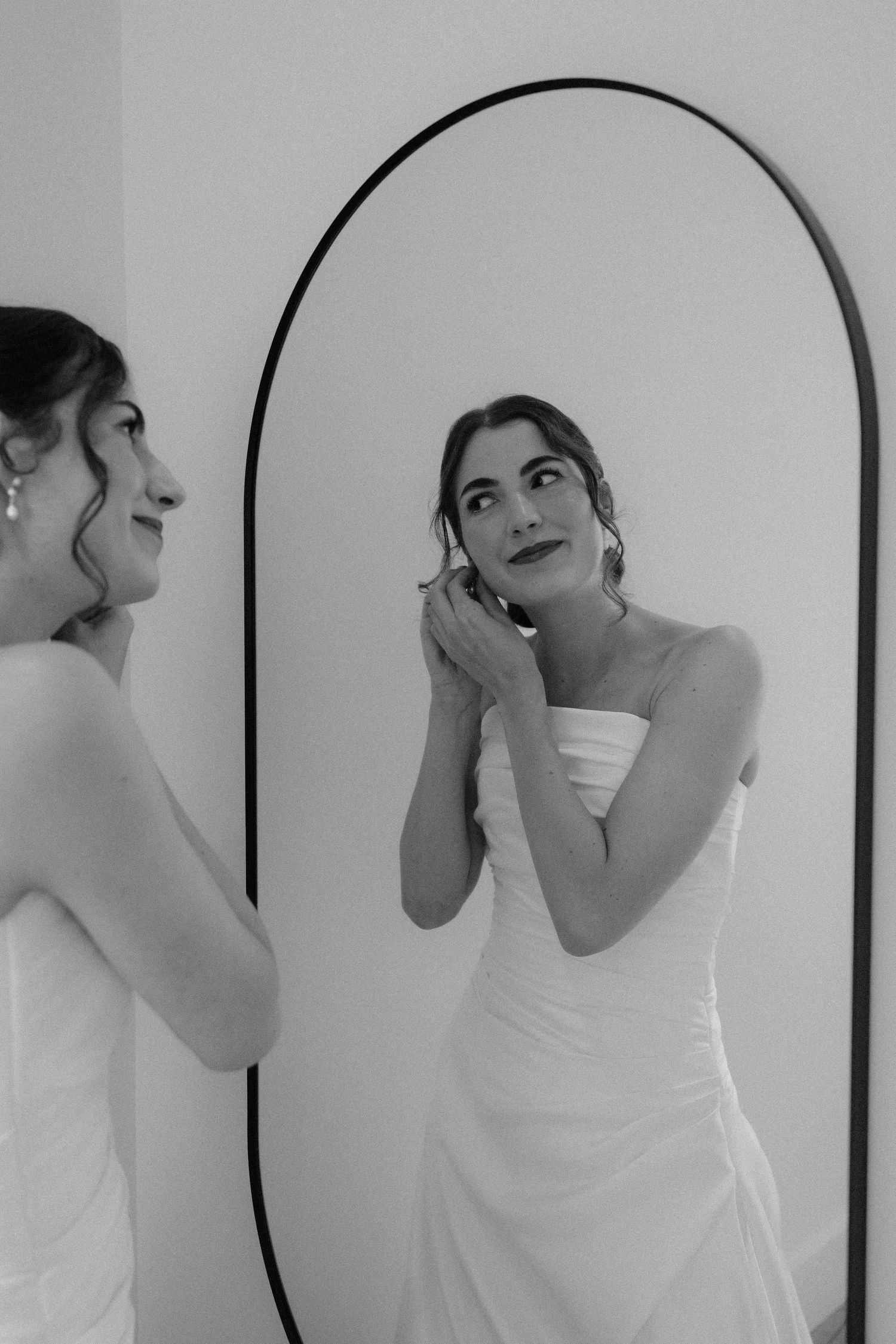 Bride in strapless white gown adjusting earring while gazing into an arched black-framed mirror, black and white photo.