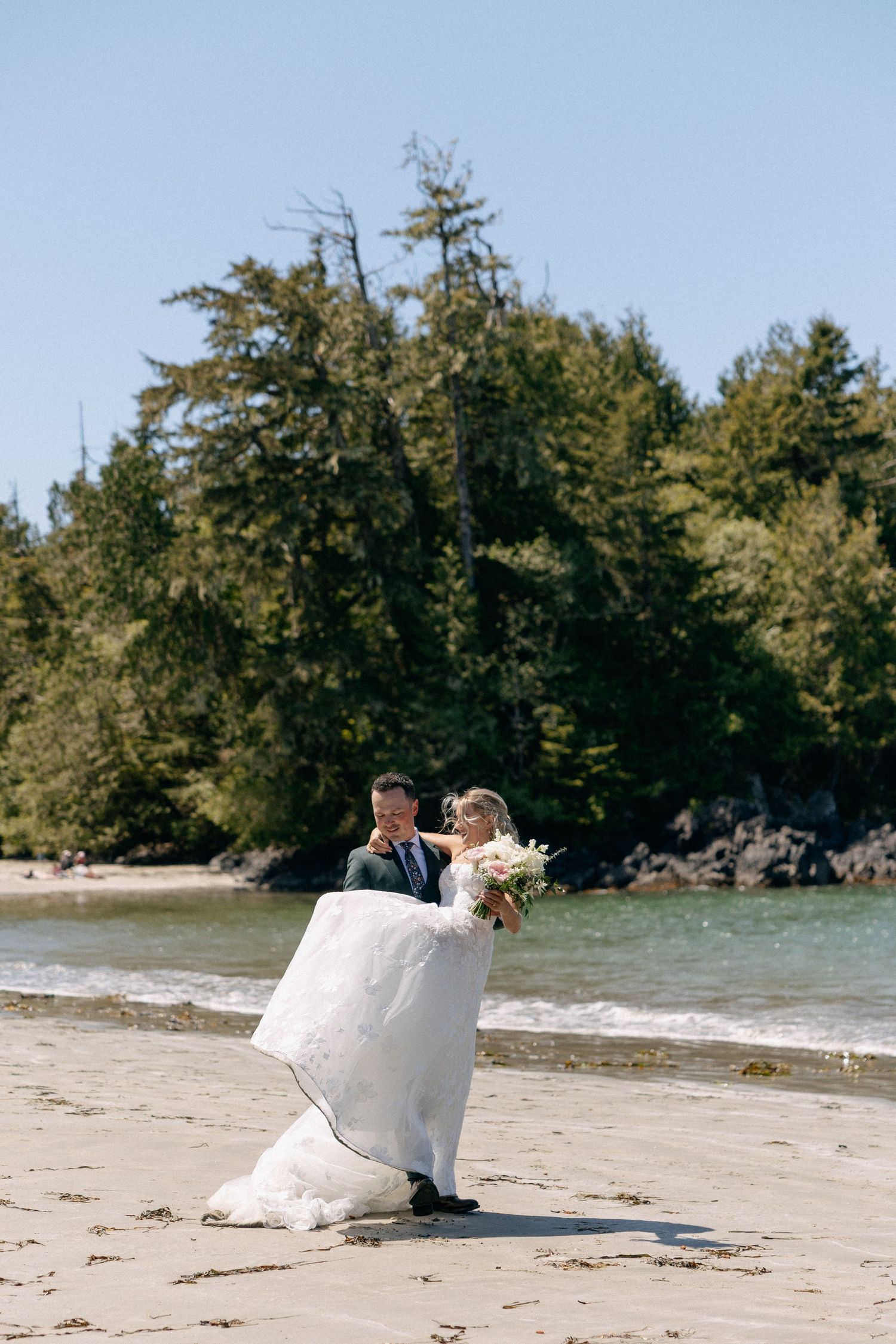 Bride and groom dancing on a sandy beach with lush green trees and ocean waves in the background on a sunny day.