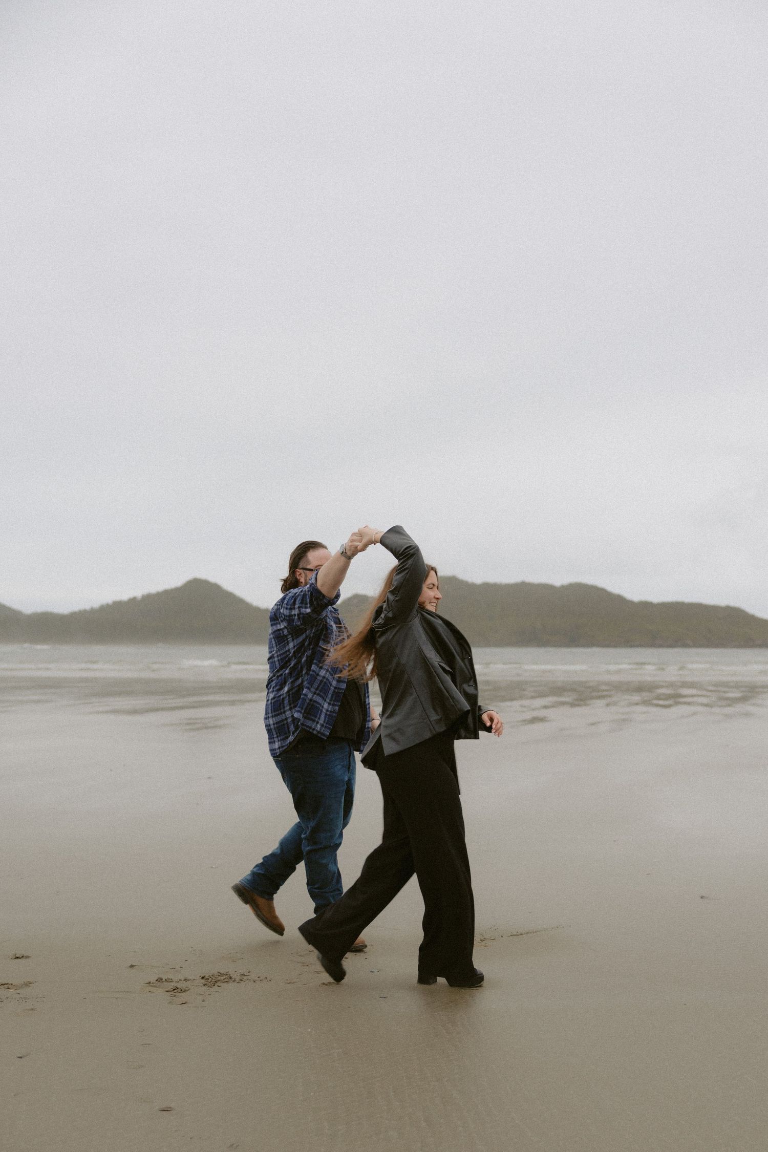 Tofino proposal couple dancing and twirling on a misty beach with mountains visible in background on Chesterman Beach