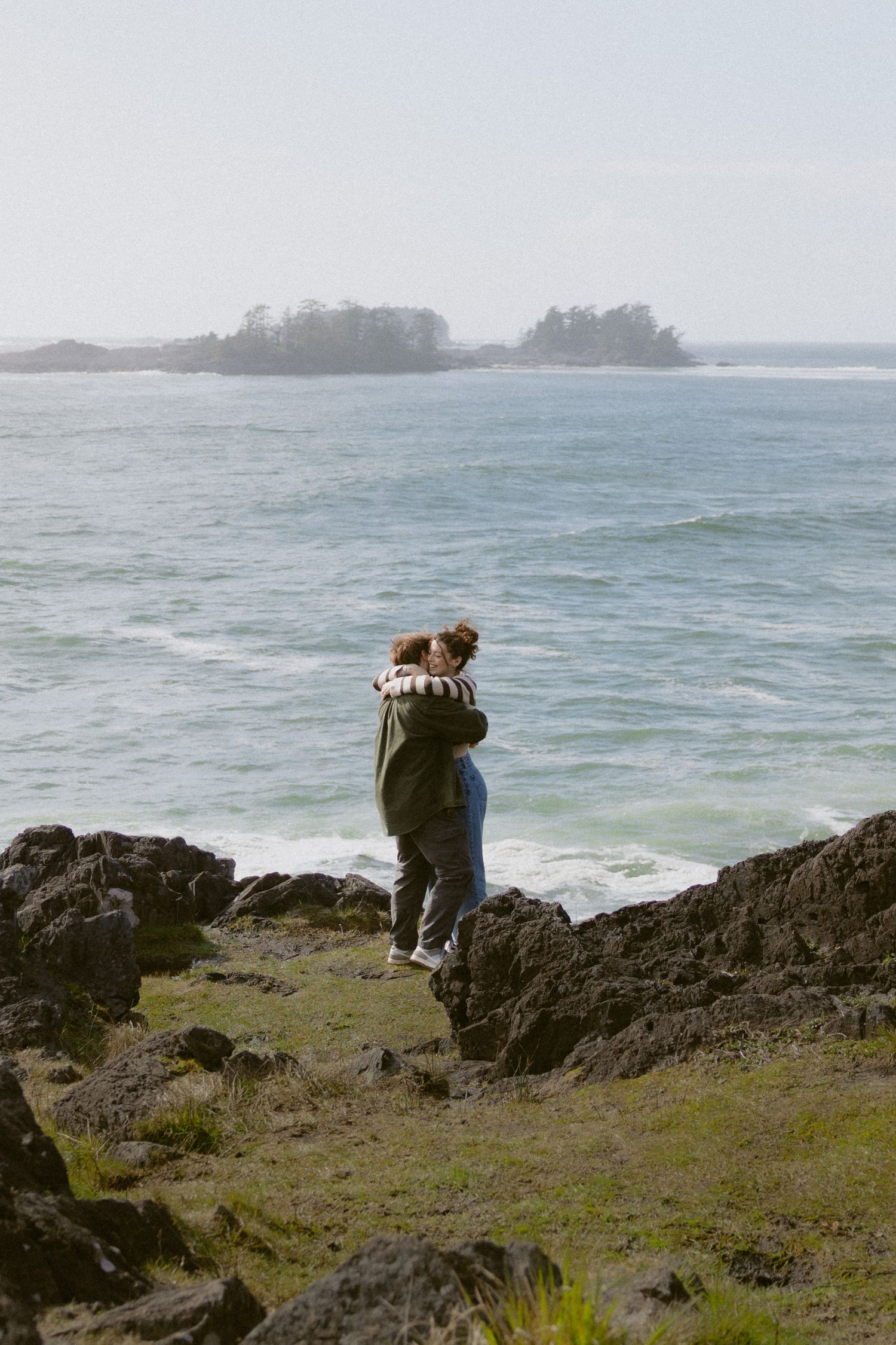 Couple hugging on mossy coastal rocks with scenic ocean island view on overcast day.