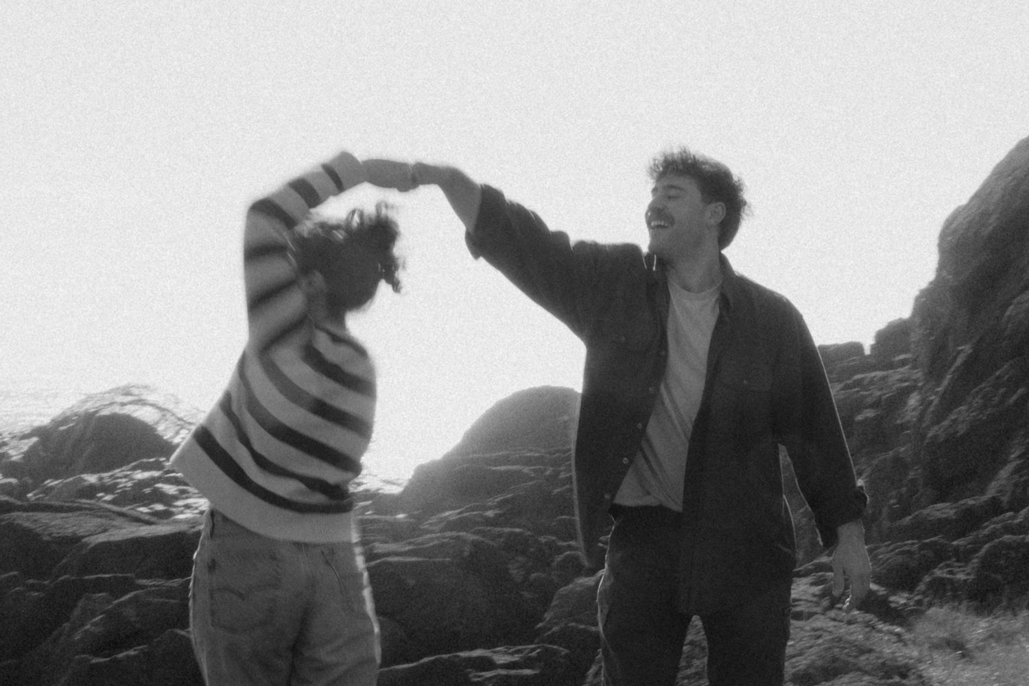 Black and white photo of playful couple dancing together among coastal rocks outdoors.