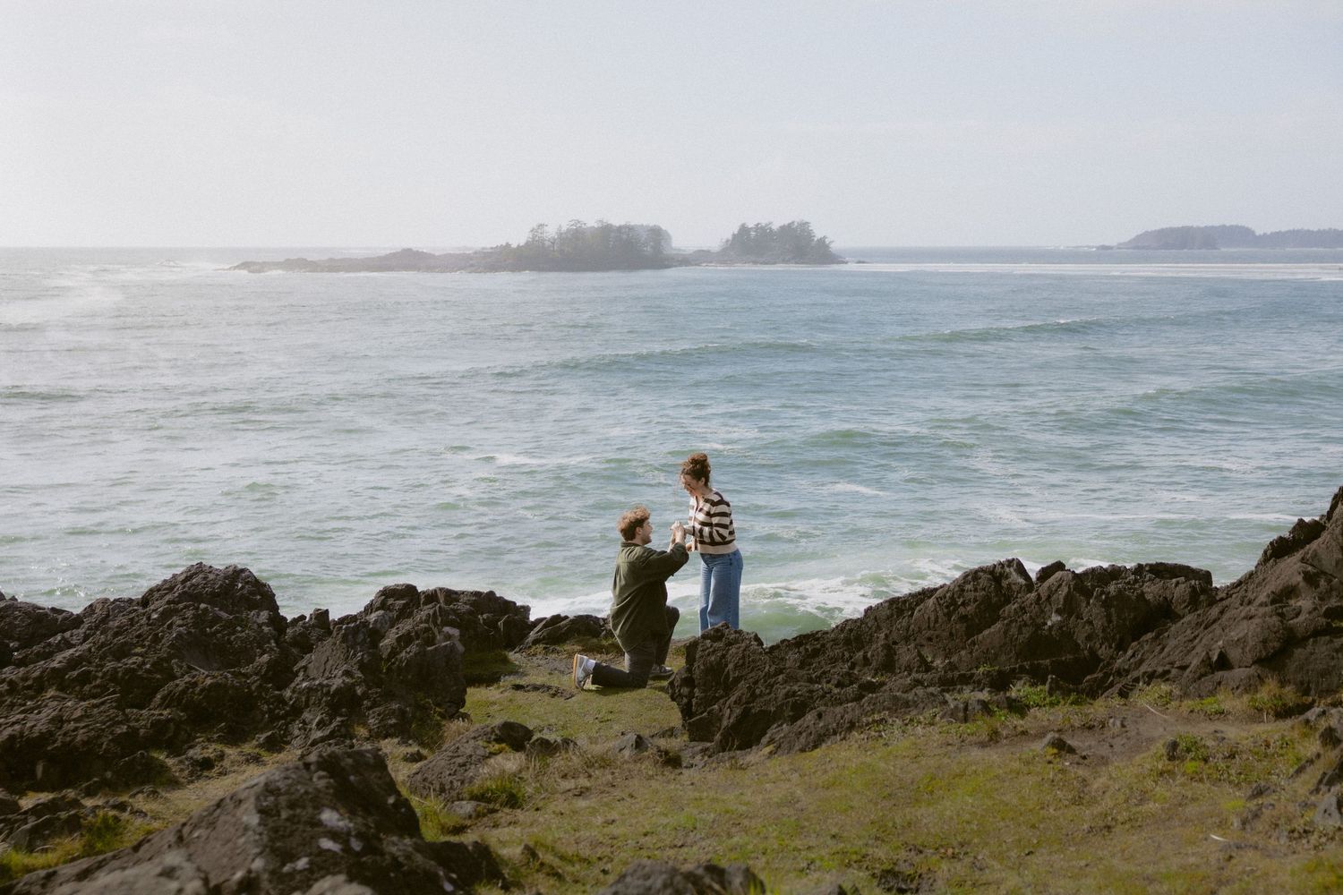 Couple on rocky coastal cliffs with ocean waves and misty islands in background during proposal moment.