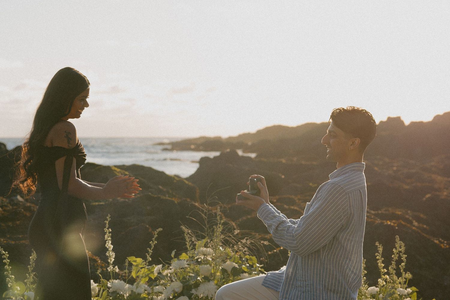 Man kneeling on rocky coastal cliff proposing to woman at golden sunset.