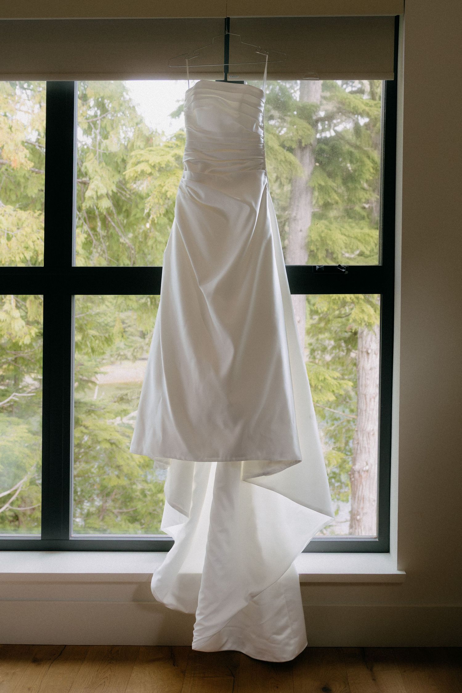 White strapless wedding gown hanging on a hanger in front of a large window with green trees outside.
