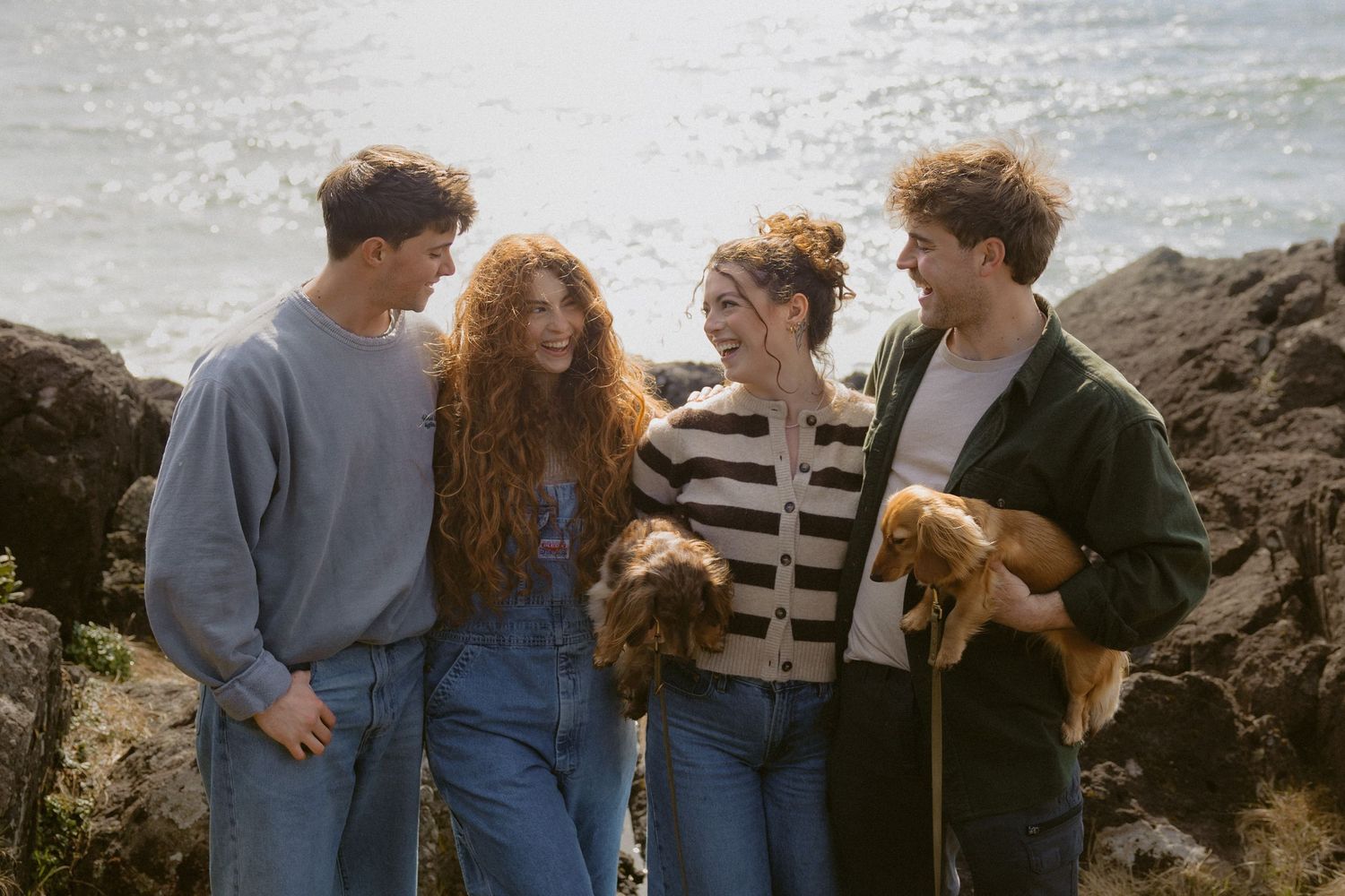 Four laughing friends holding golden dogs on sun-drenched rocky coastal cliffs with sparkling ocean behind them.