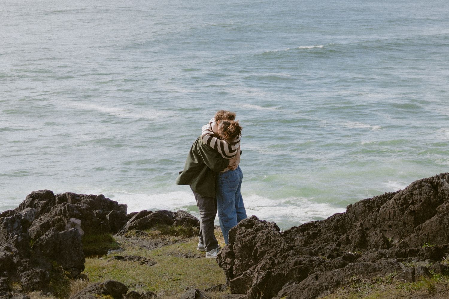 Romantic couple embracing and kissing on rocky shoreline with turquoise ocean waves behind them.