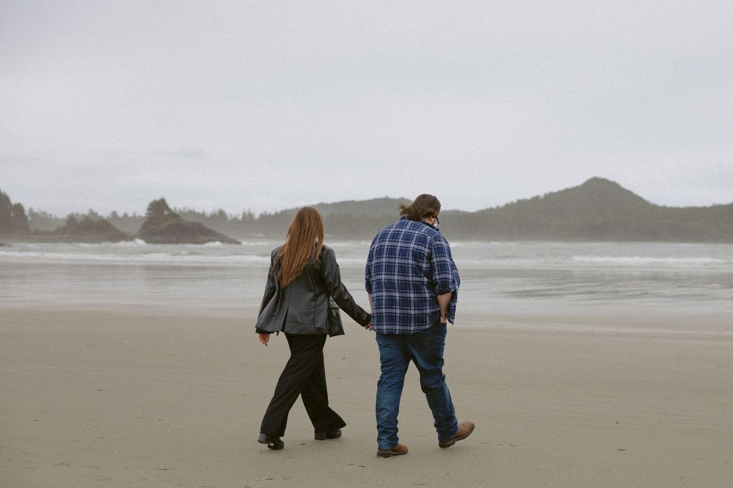 Two people walking along a misty beach shoreline with mountains in the distance.