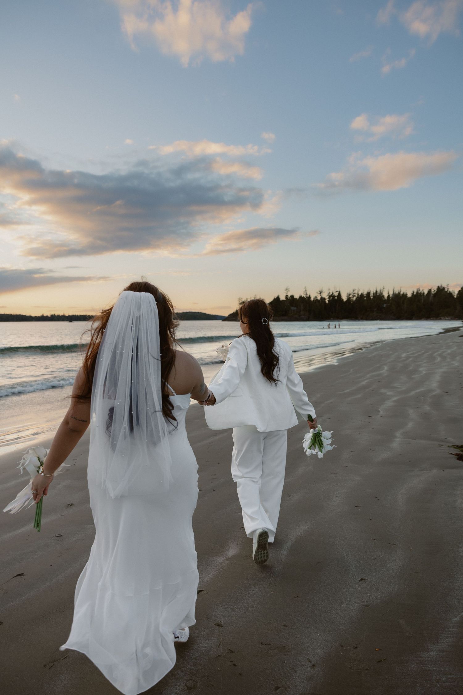 Two people in white outfits walk hand in hand along the shoreline during sunset.
